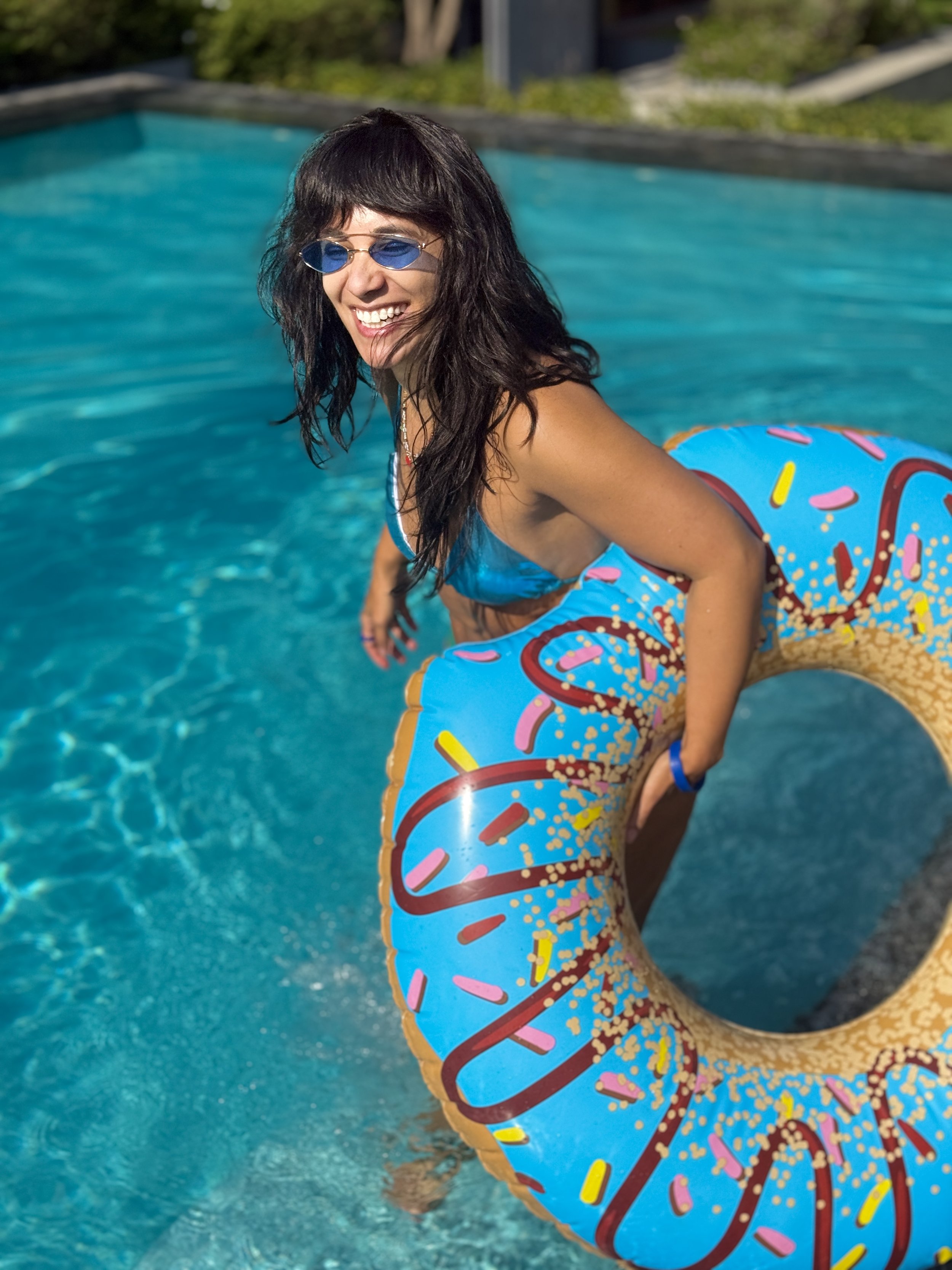 A woman with dark hair and sunglasses smiling while sitting on a donut-shaped pool float in a swimming pool.