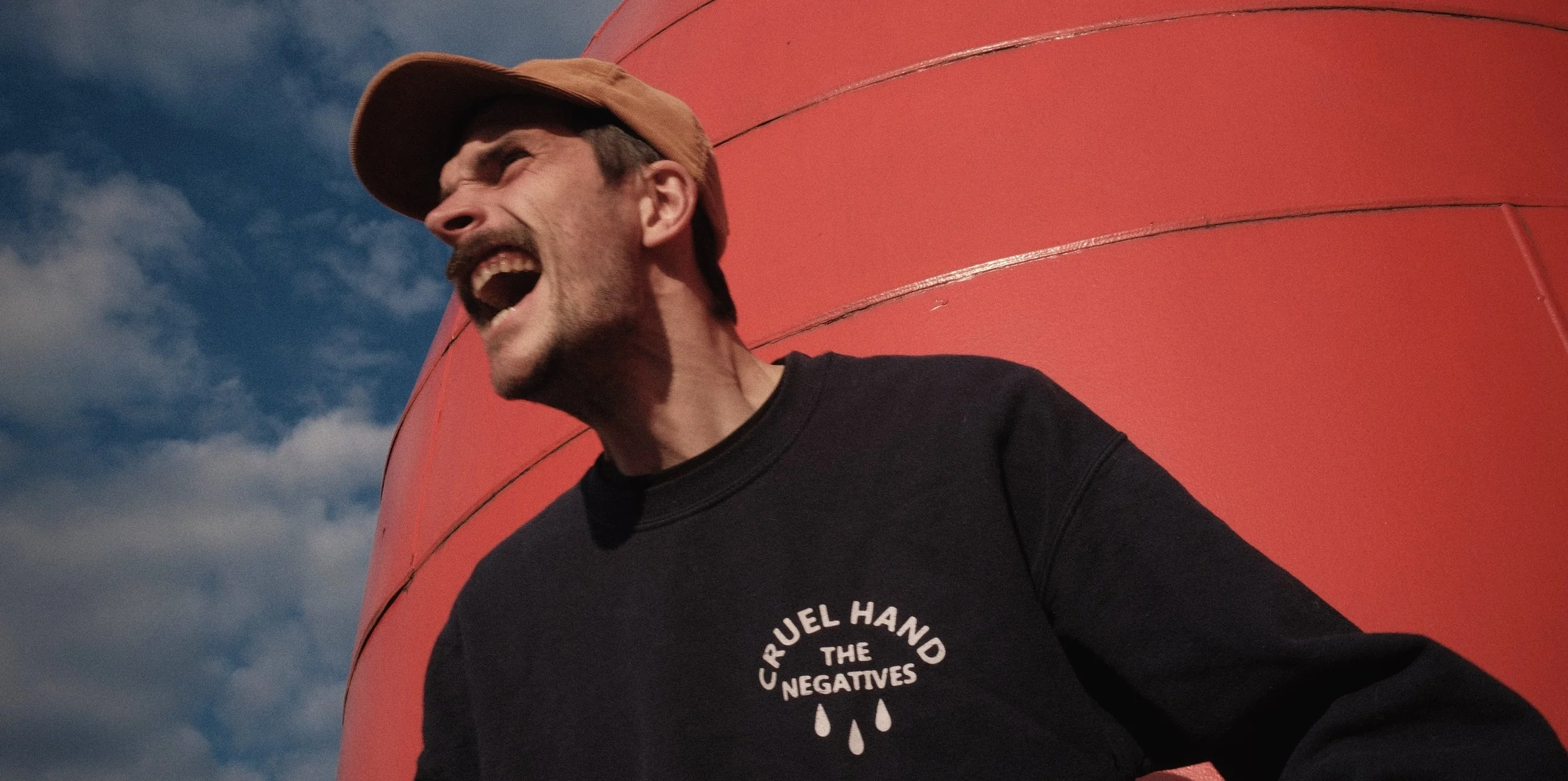Ein Mann schreit lachend neben einem großen roten Objekt, trägt ein schwarzes T-Shirt mit weißem Text und einem Tropfensymbol, bei bewölktem Himmel.