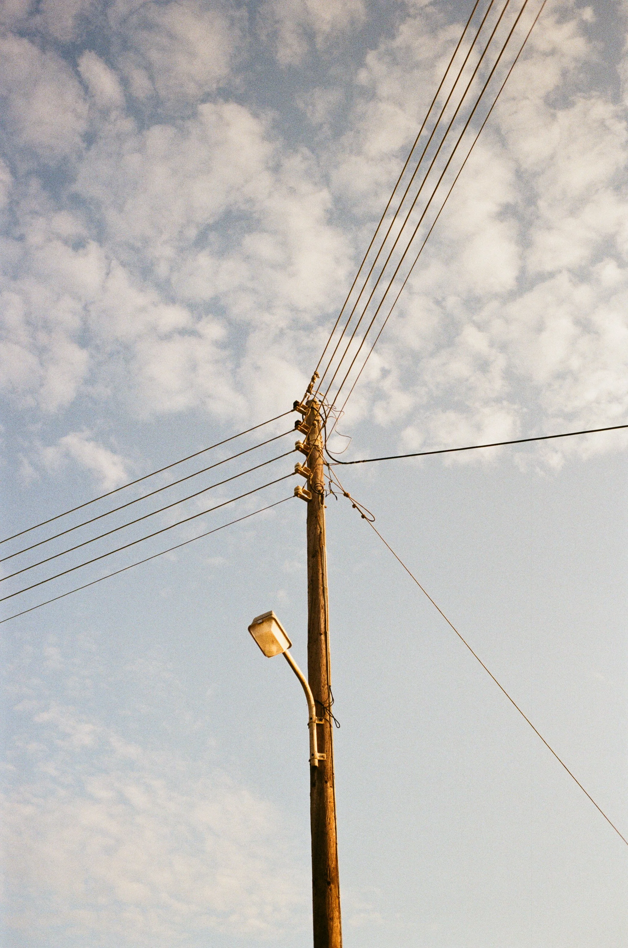 Holzmast mit mehreren Stromleitungen und einer Straßenlaterne gegen den Himmel mit Wolken