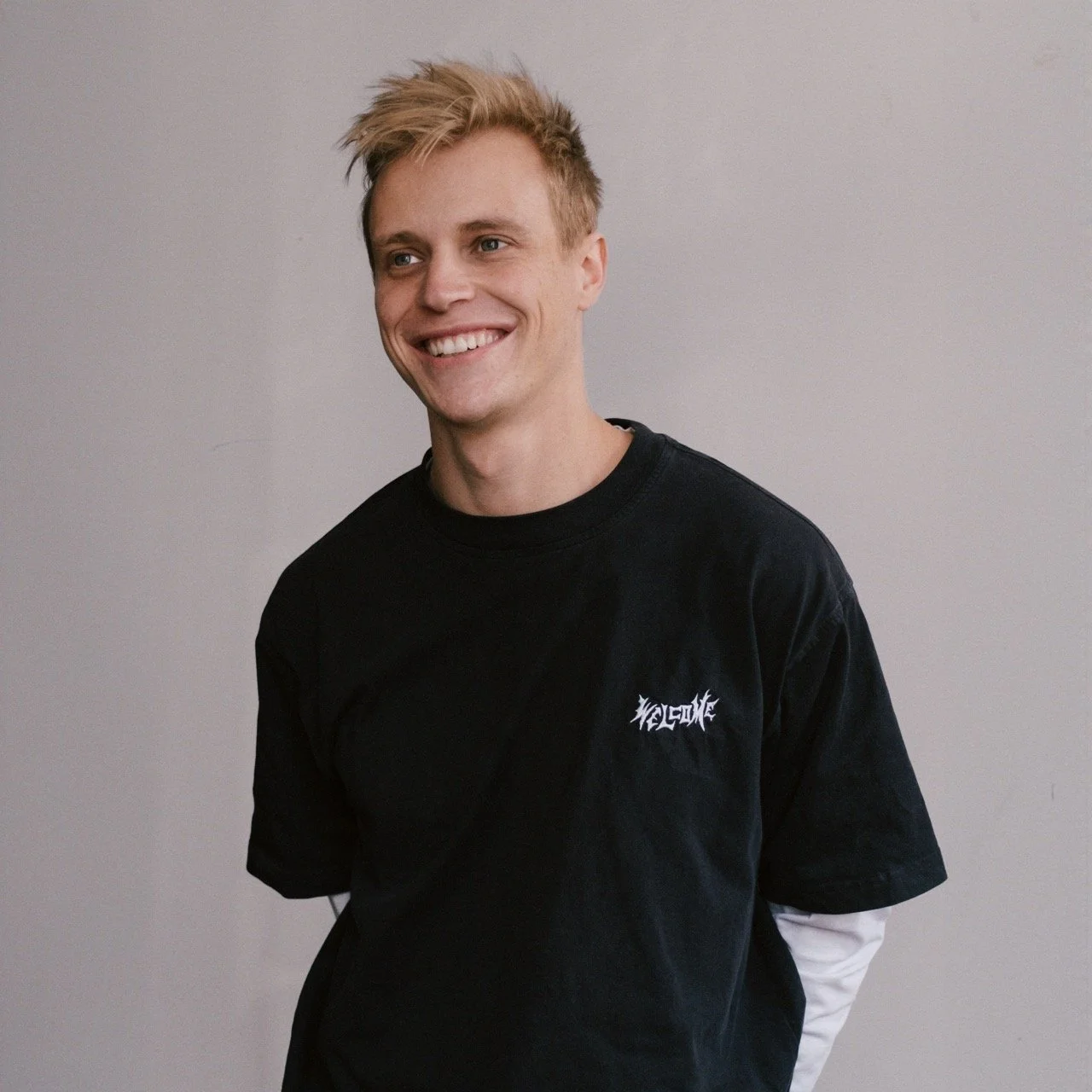 Ein lachender junger Mann mit blondem, zerzaustem Haar trägt ein schwarzes T-Shirt mit einem weißen Logo auf der Brust, steht vor einer hellgrauen Wand.
