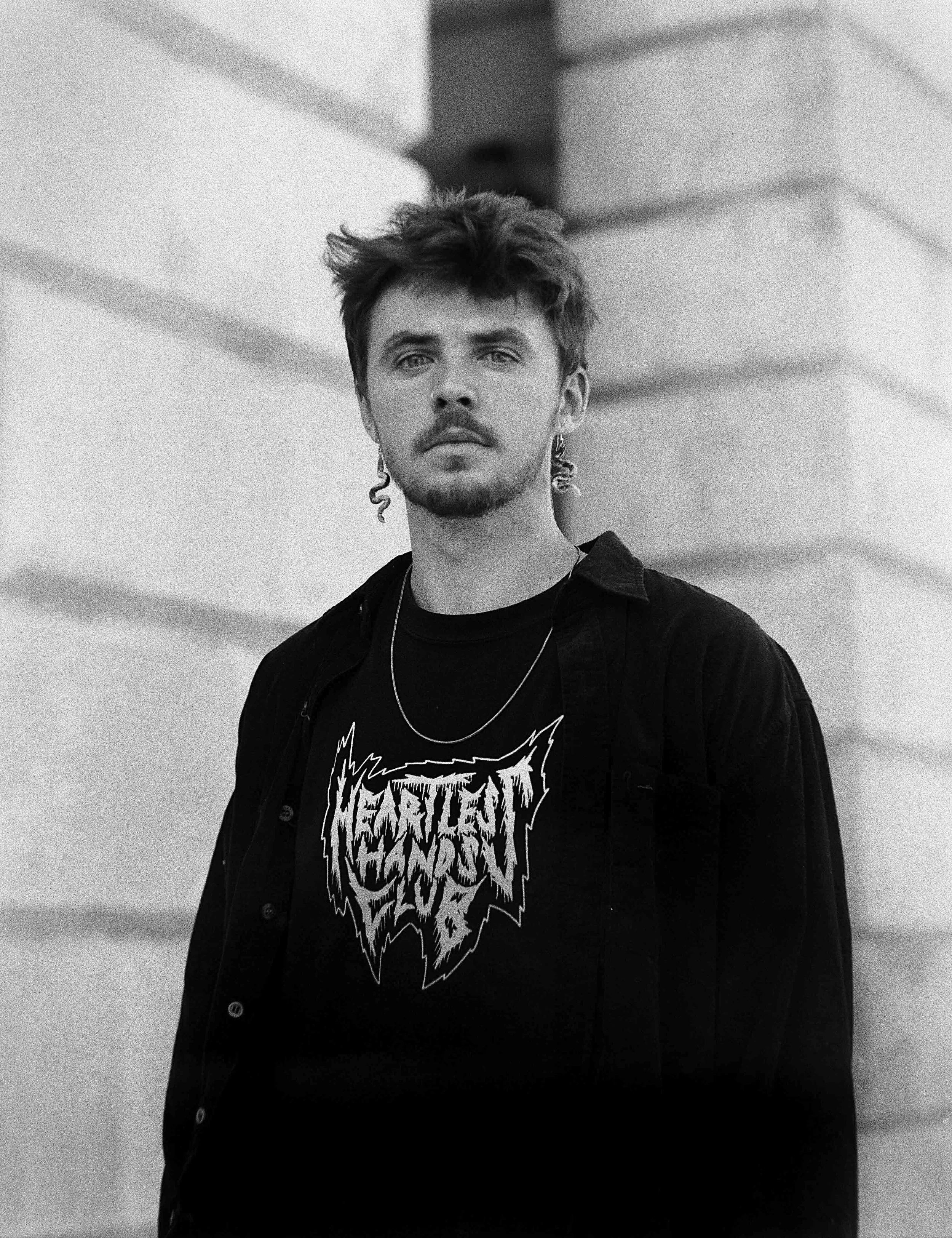 Schwarz-Weiß-Fotografie eines jungen Mannes mit zersausten Haaren, Bart und Eidechsenohrringen, der ein T-Shirt mit dem Schriftzug 'Metallica' trägt, vor einem modernen, schrägen Gebäudearchitektur-Hintergrund.
