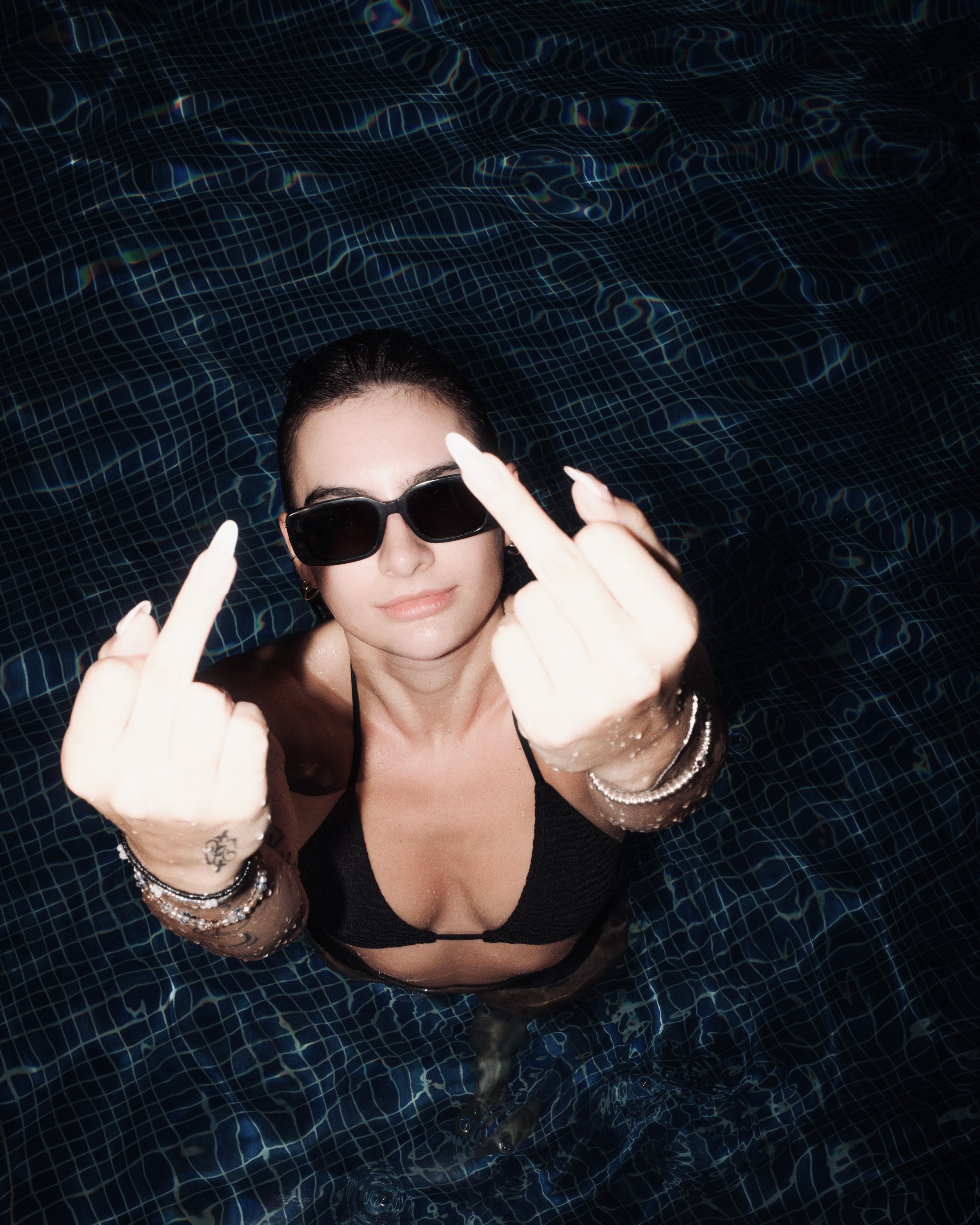 Frau mit Sonnenbrille und Schmuck, im Wasser liegend, zeigt mit beiden Händen die Mittelfinger, umarmt eine Meerjungfrau im Pool.