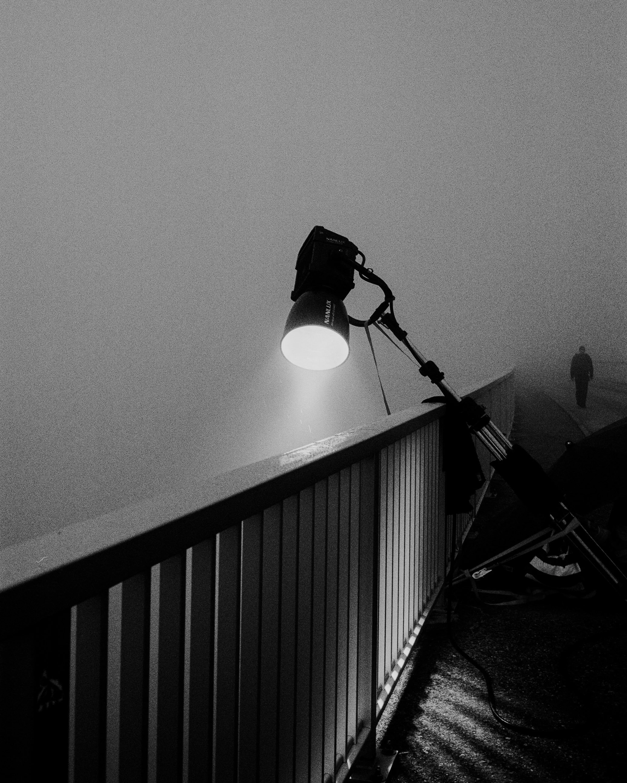 Ein Laternenstrahler leuchtet hell in eine dunkle, dämmerige Umgebung. Im Hintergrund sind Silhouetten von Gebäuden und einer Person sichtbar.