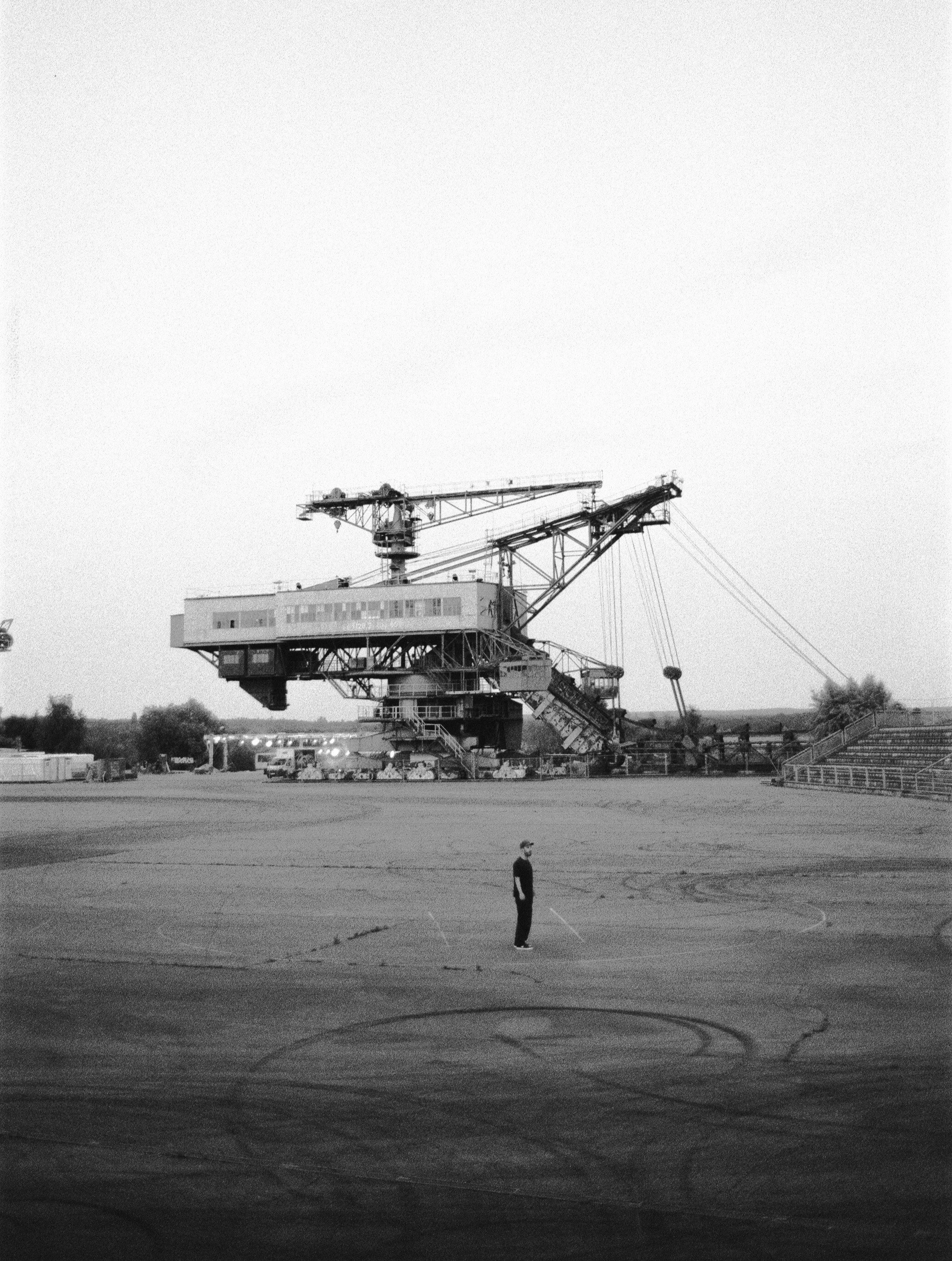 Schwarz-Weiß-Foto eines Mannes, der auf einem leeren Flugfeld steht, im Hintergrund eine große Baumaschine