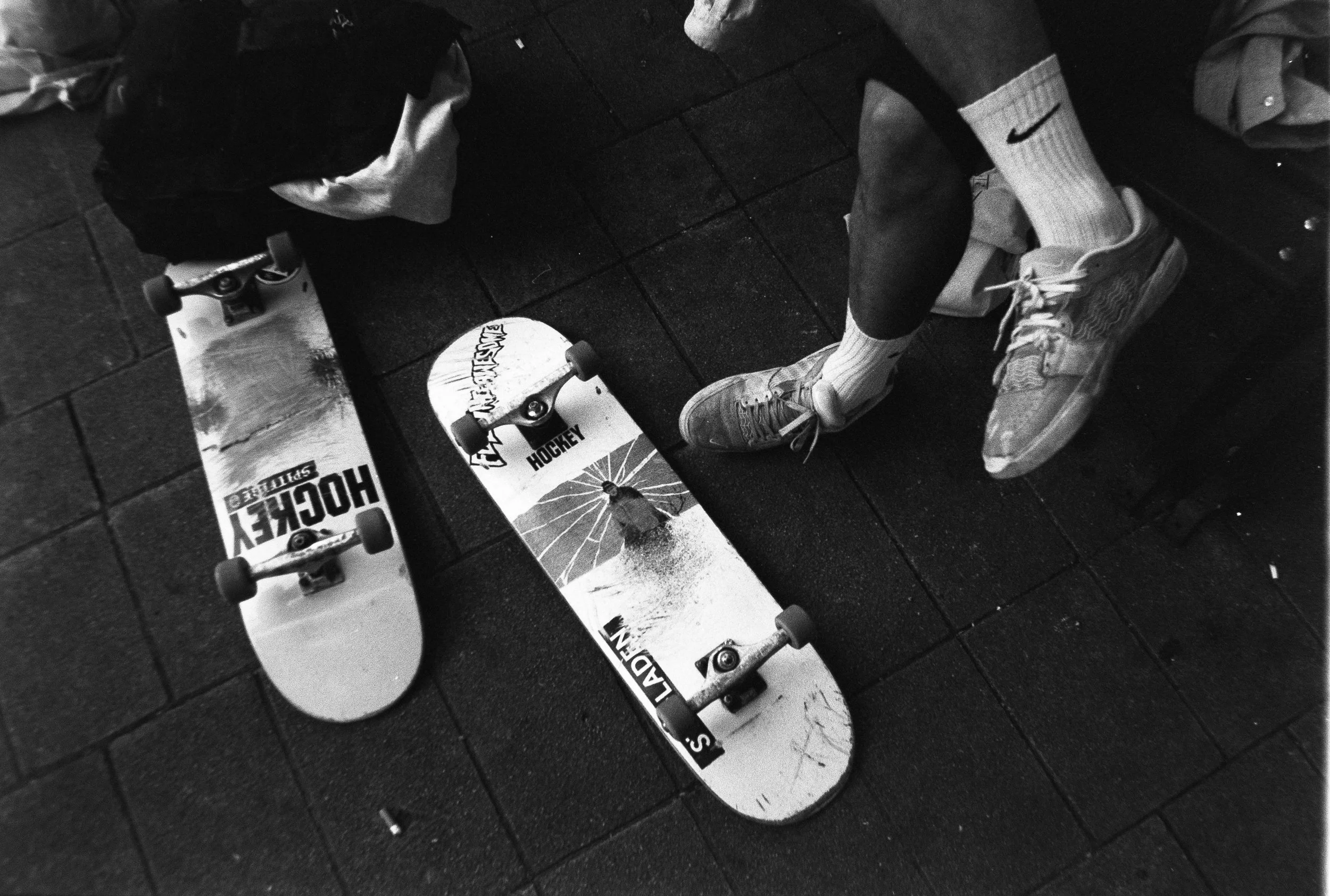 Schwarz-weiße Aufnahme eines Skateboards auf einem Boden, umgeben von einem Kind, das Sportschuhe und Socken trägt, und einer Person in einem Mantel.