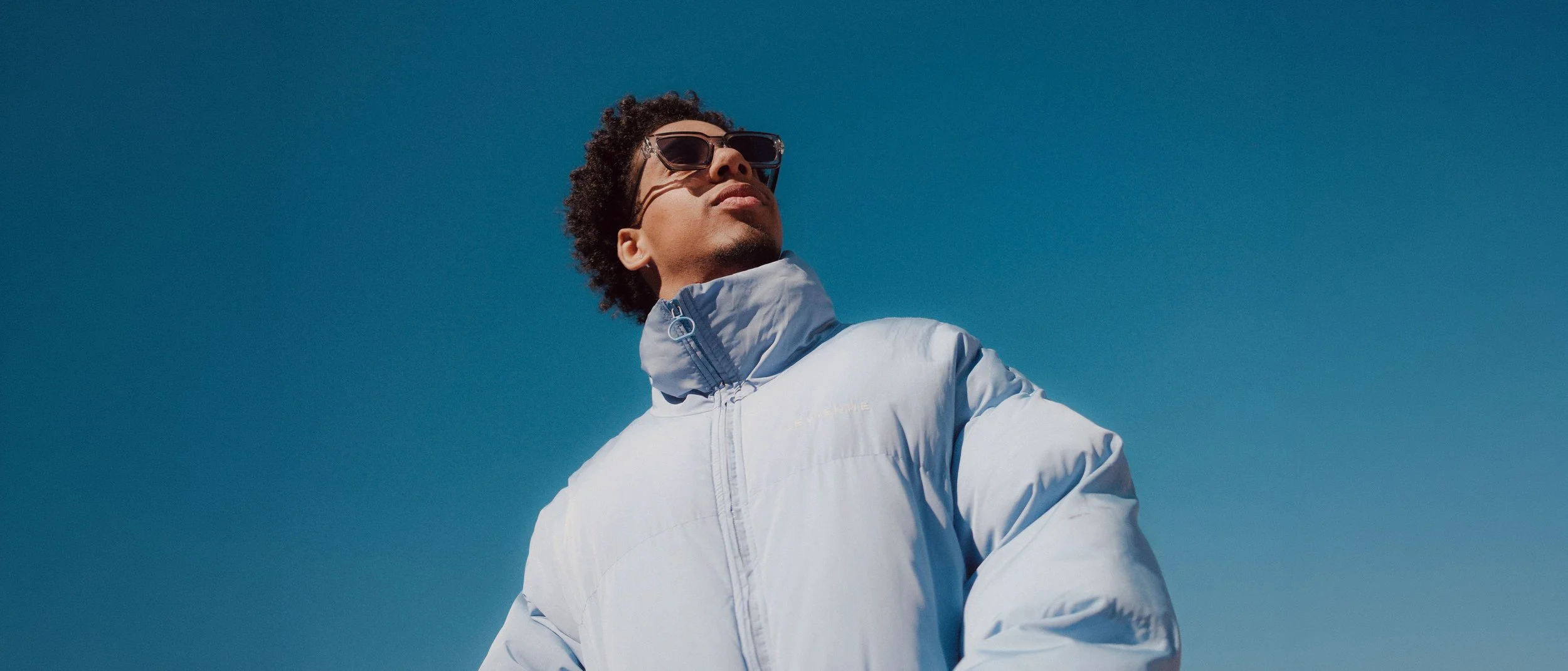 Junge Person trägt eine weiße Jacke und Sonnenbrille, blickt in die Ferne, blauer Himmel.