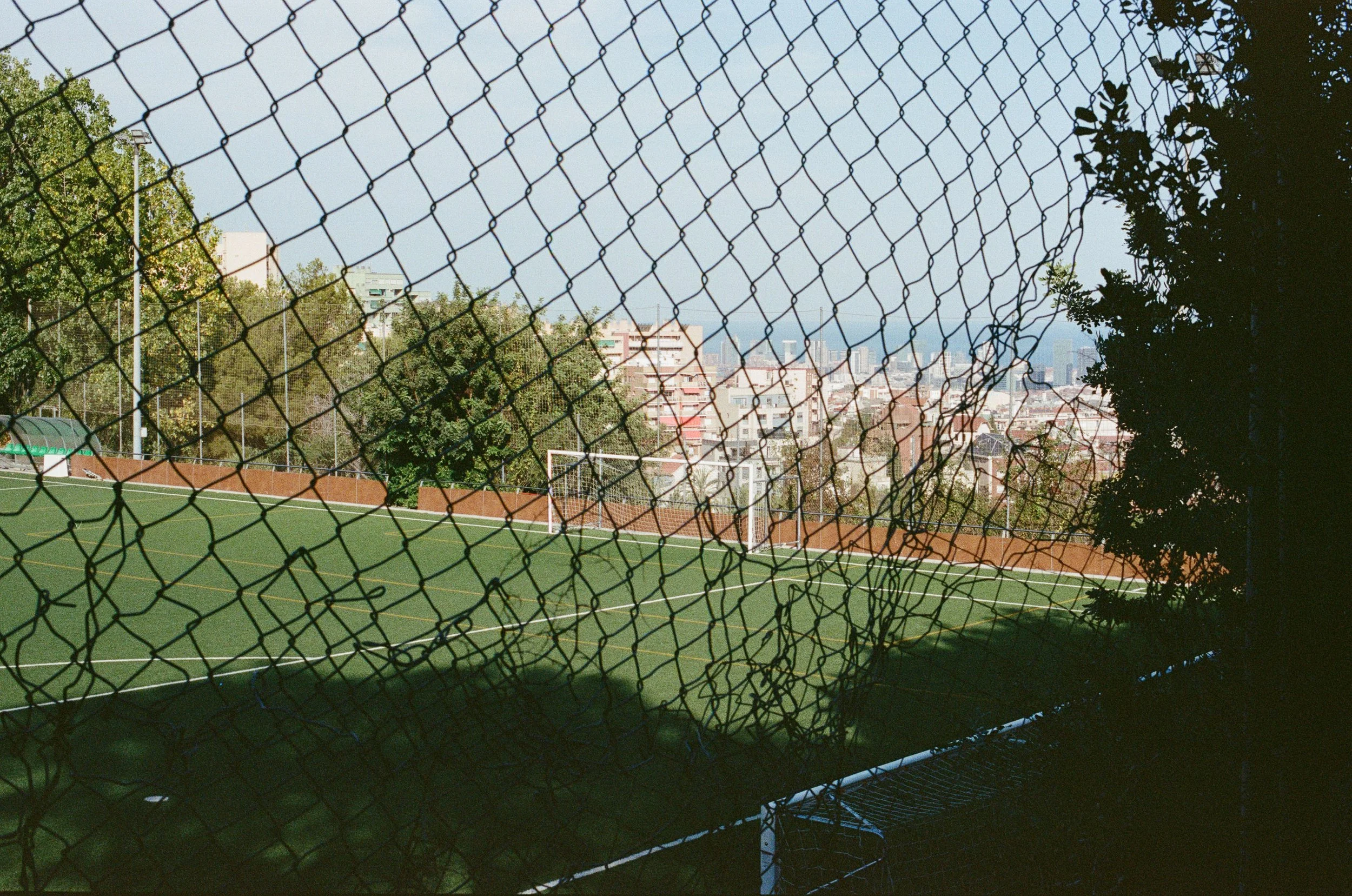 Ein Sportplatz mit Tor und Umzäunung, vor Hintergrund eine Stadt mit Gebäuden und Bäumen, Blick durch einen Zaun hindurch.