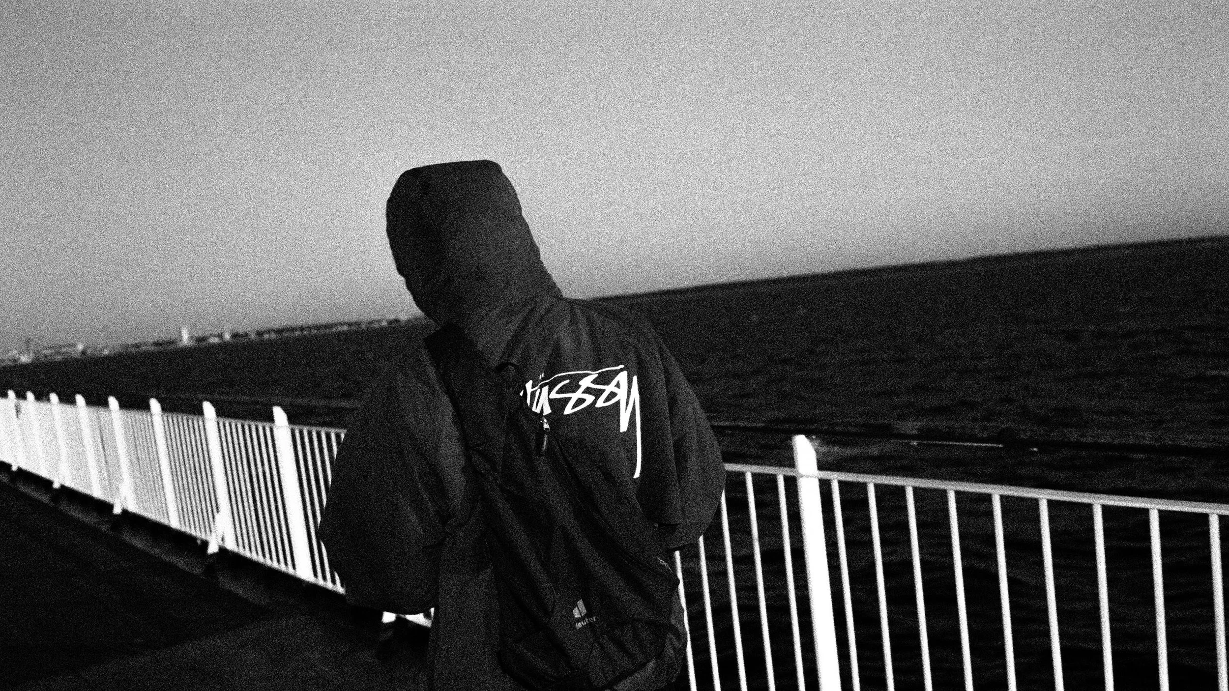 Eine Person mit Kapuzenjacke steht an einem Geländer am Meer bei Nacht, Blick auf den Horizont, im Schwarz-Weiß-Foto.