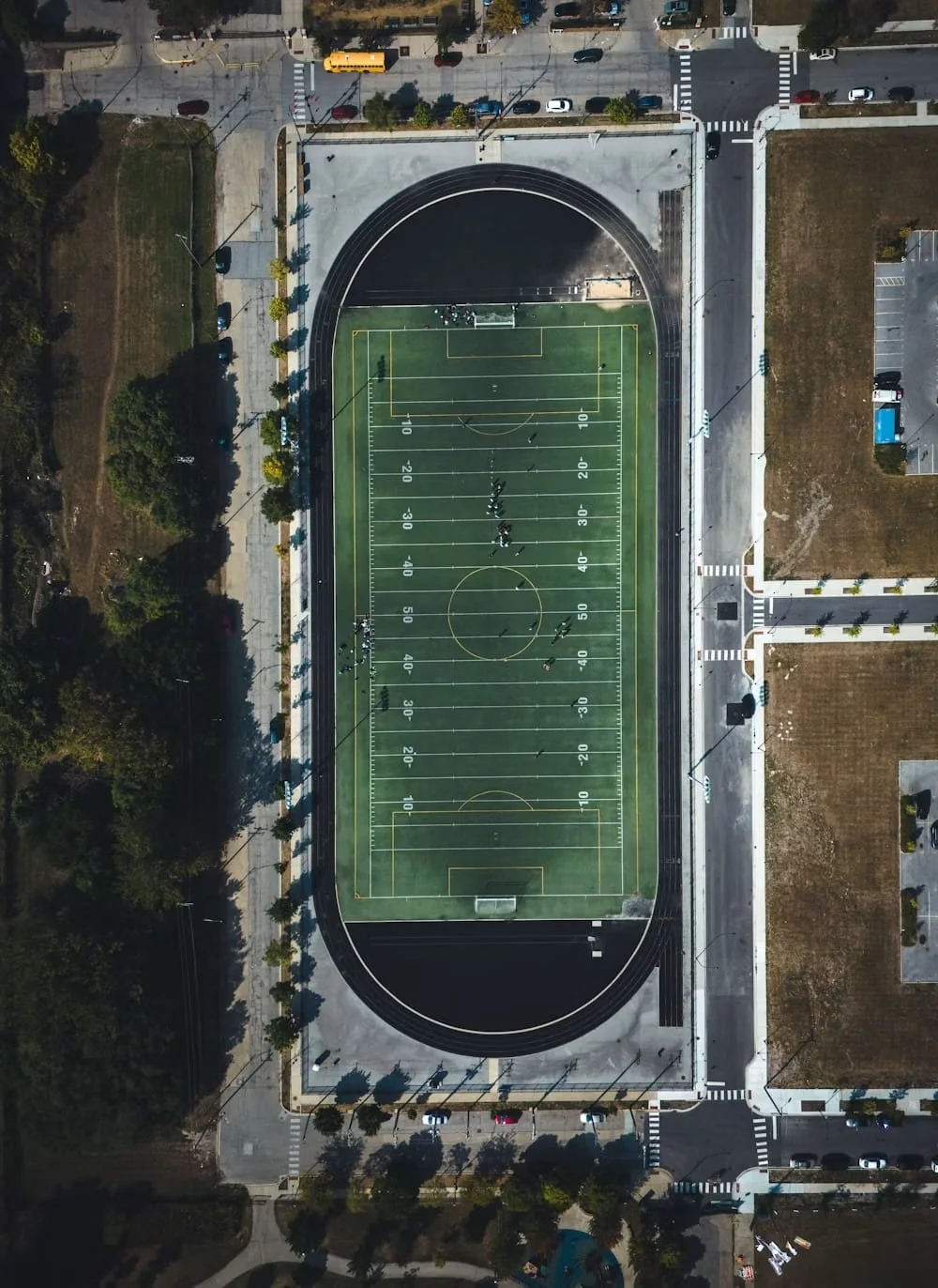 Aerial view of a high school football field with a running track around it, surrounded by parking lots, streets, trees, and nearby buildings.