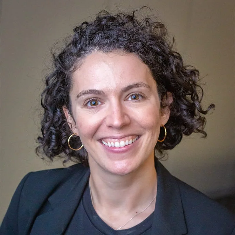 A woman with curly dark hair smiling, wearing gold hoop earrings, a black blazer, and a black top, against a plain background.