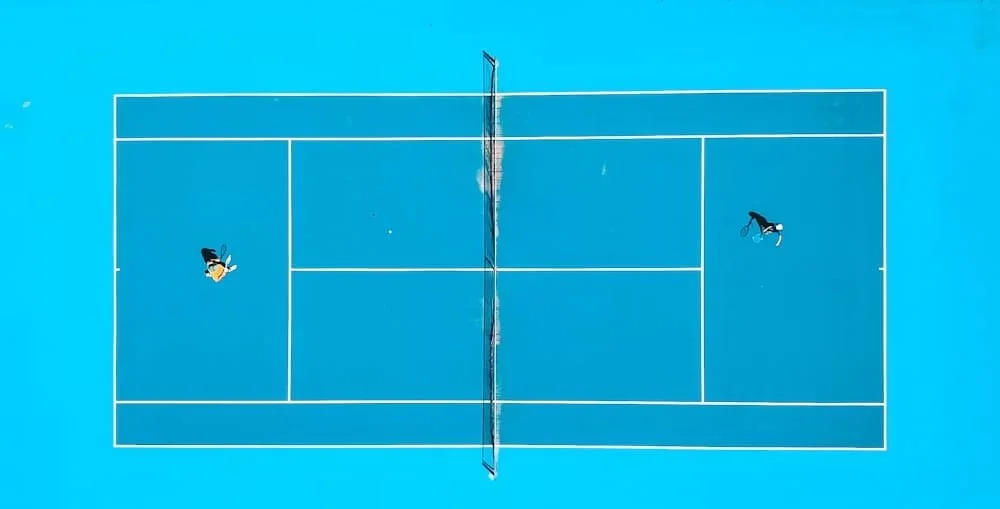 Overhead view of a blue tennis court with a net in the center. Two cartoon characters, one on each side of the net, are playing tennis.