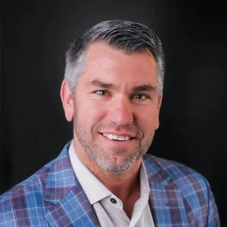 Portrait of a middle-aged man with short gray hair and a beard, smiling, wearing a blue plaid blazer and white shirt against a black background.