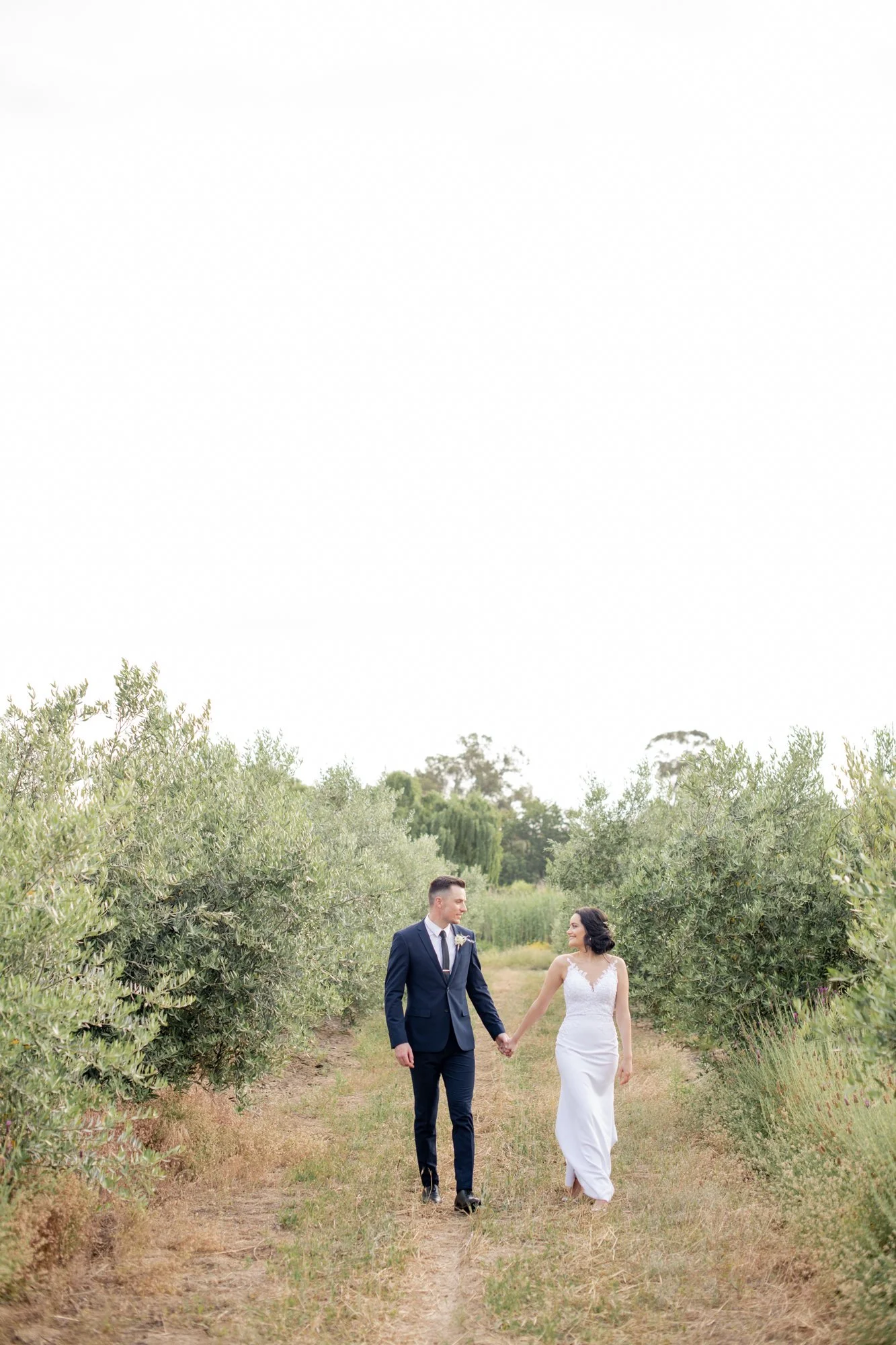 A bride and groom holding hands and walking down a dirt path surrounded by green bushes and trees in a natural outdoor setting.