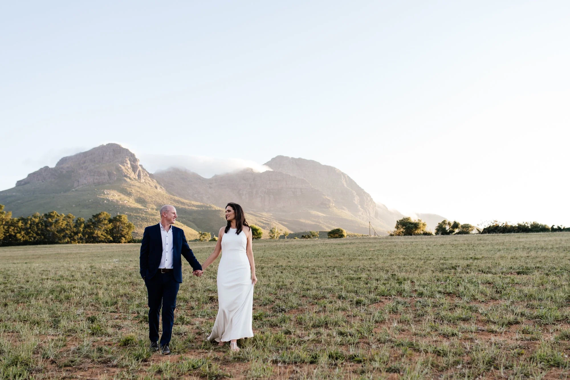 A couple holding hands, walking in an open grassy field with mountains in the background during daylight.