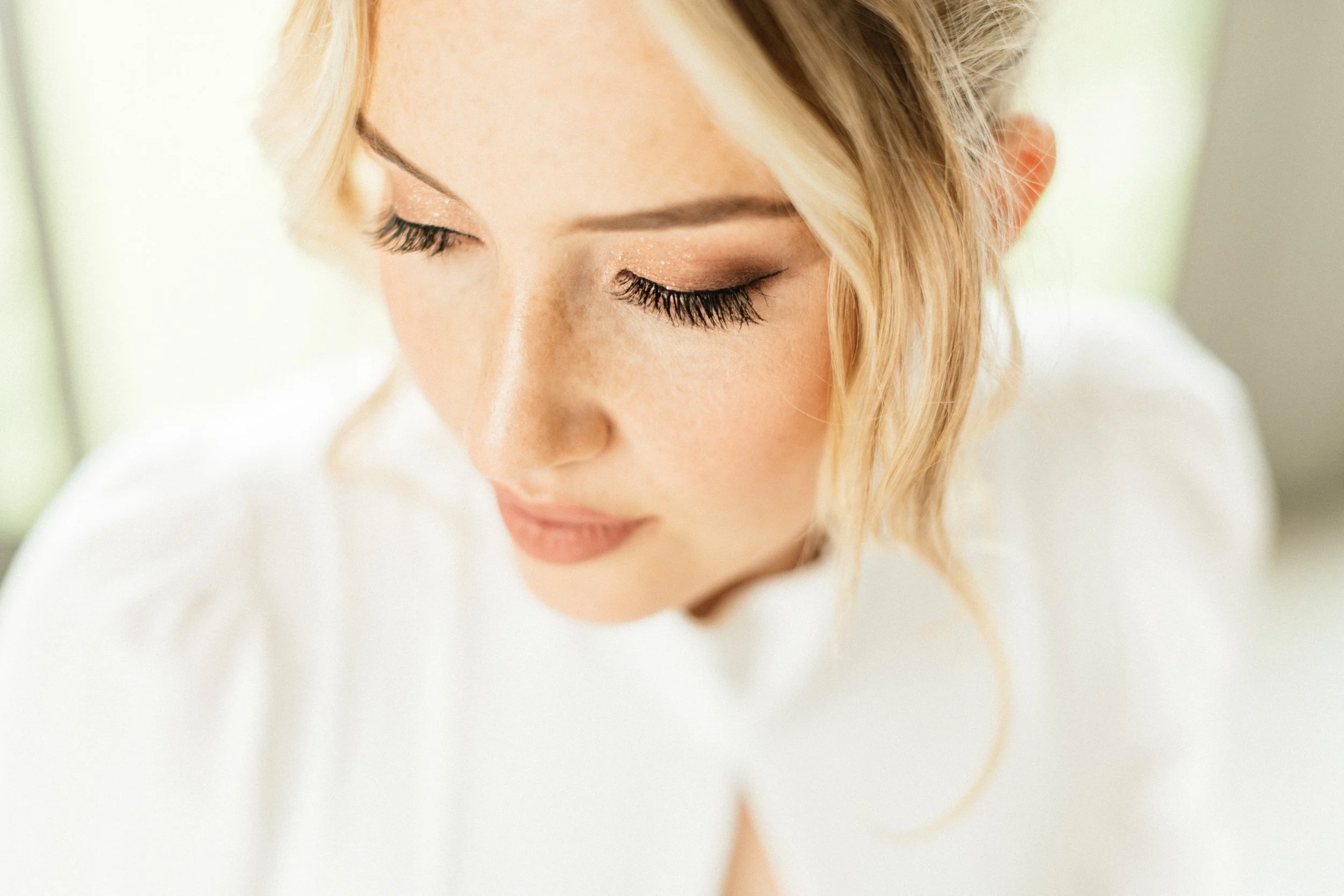 Close-up of a woman with blonde hair and closed eyes, wearing light makeup with glittery eyeshadow and long eyelashes.