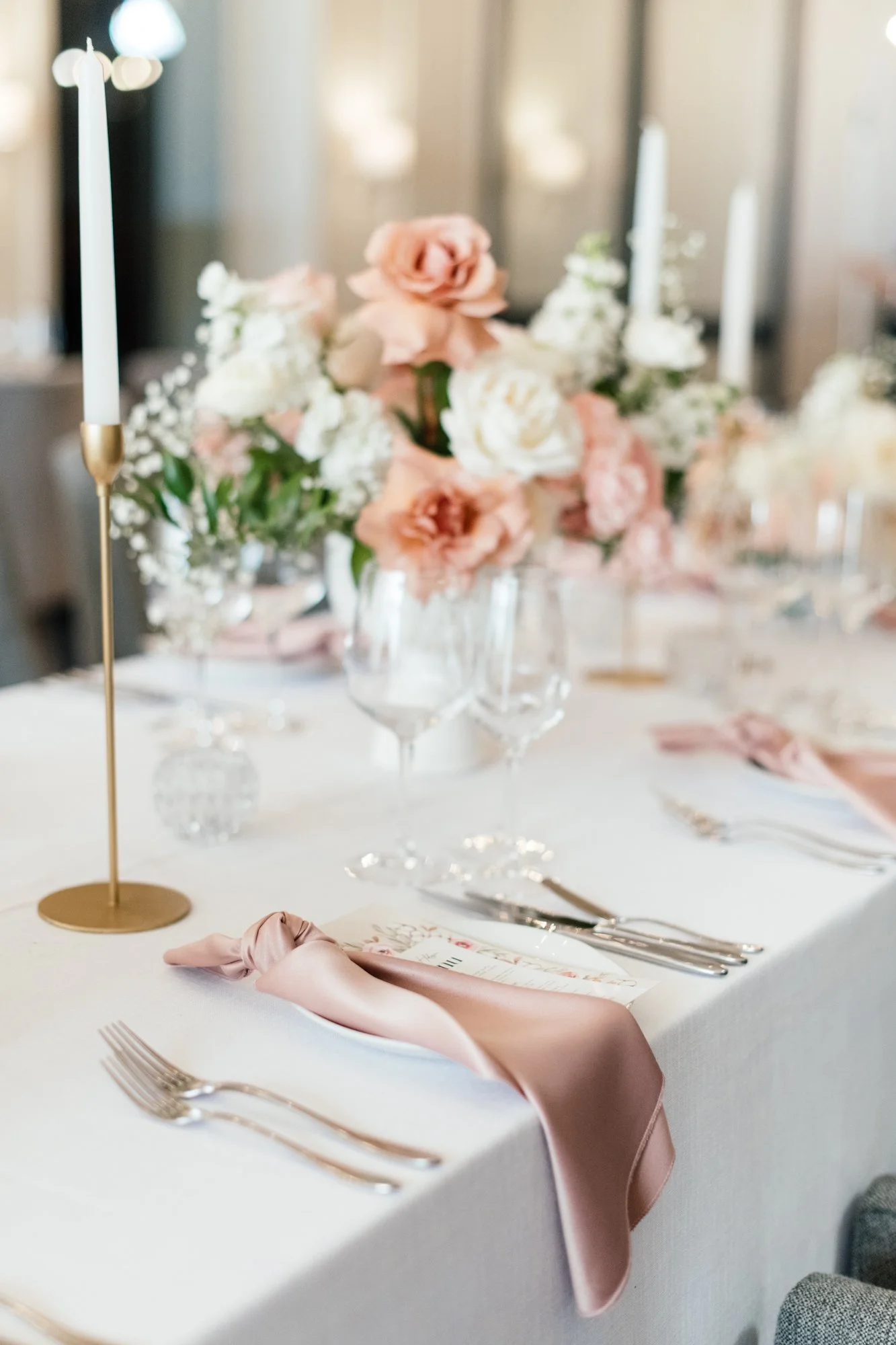 A wedding reception table set with a white tablecloth, a pink satin napkin, silverware, and glassware. There is a floral centerpiece with white and pale pink flowers, and a gold candle holder with a white candle. The background shows a softly lit roo
