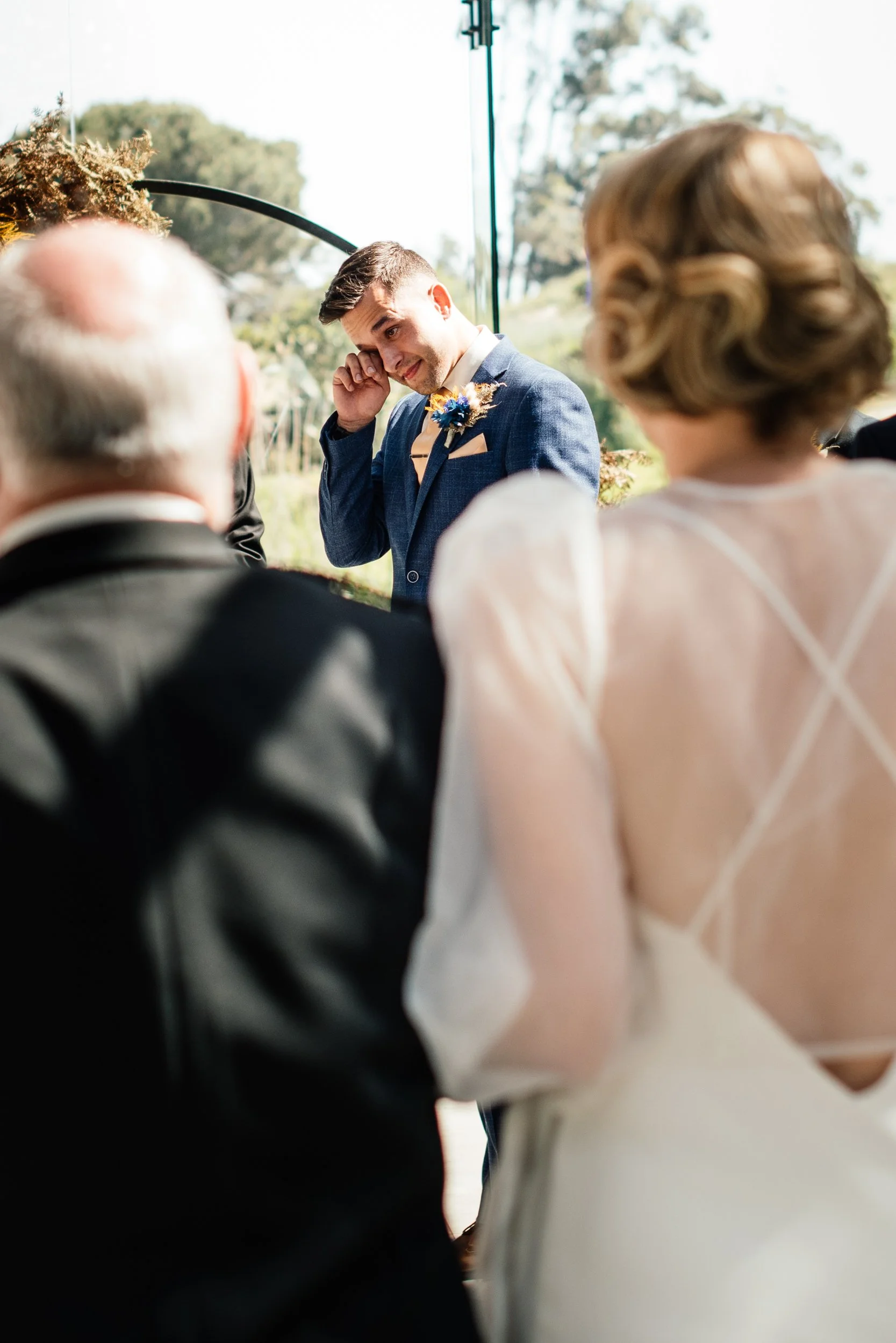 A man in a blue suit is wiping tears from his eyes during a wedding ceremony, with older and younger women in the foreground watching. The event is outdoors with trees and a glass wall in the background.