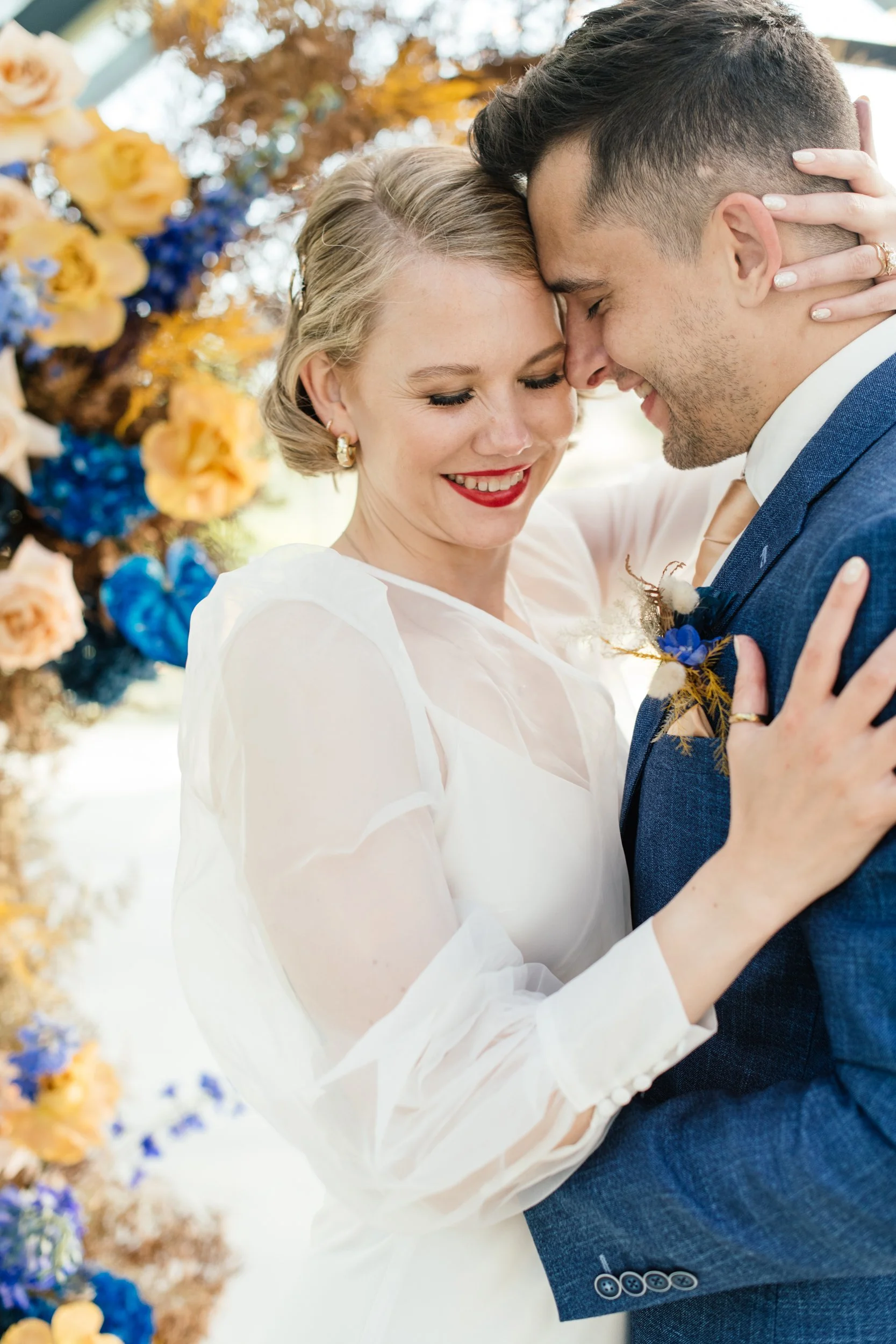 A bride and groom are embracing and smiling with their foreheads touching in a wedding scene, with a floral backdrop of yellow, blue, and brown flowers.