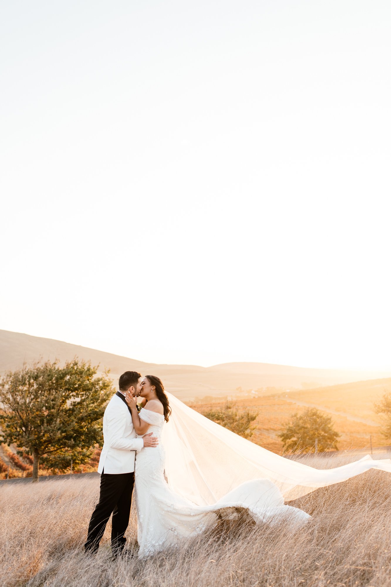 A bride and groom kiss in a field during sunset, with a white wedding dress flowing in the breeze and rolling hills in the background.