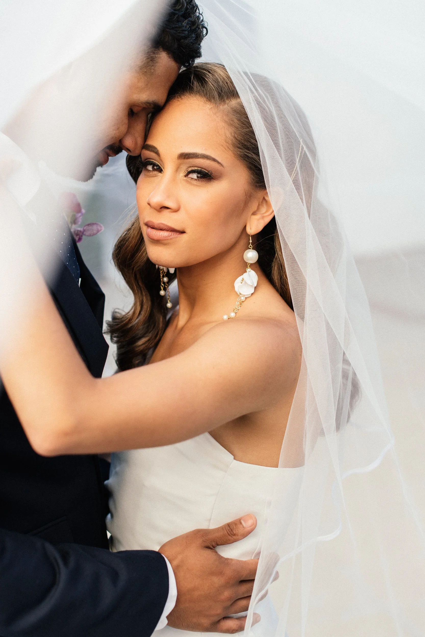 A couple on their wedding day, the groom with dark curly hair in a navy suit and the bride with long brown hair in a white wedding dress, with the groom gently holding the bride's waist and forehead touching her temple.