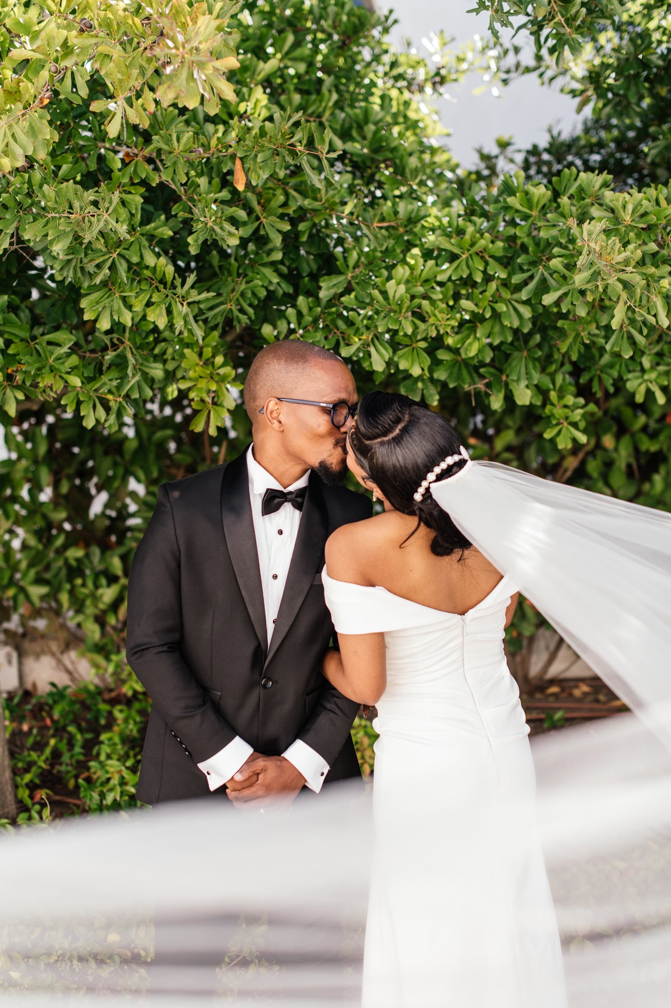 A groom in a tuxedo and bow tie kissing a bride on the forehead. The bride is wearing a white off-shoulder wedding gown and a pearl-embellished veil. They are standing outdoors in front of green bushes.