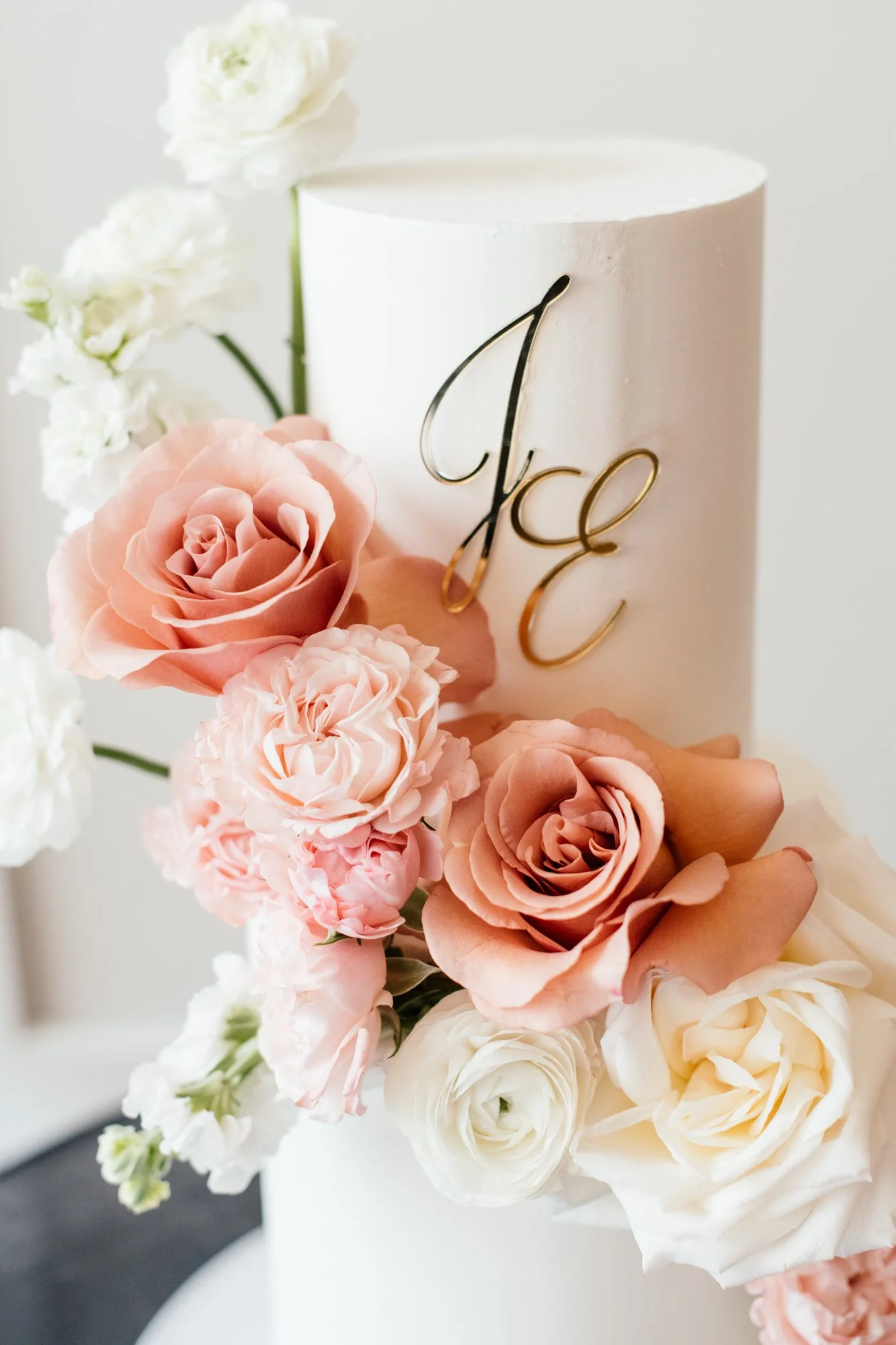 A white cake with a gold decorative monogram, surrounded by pink and white roses and white flowers.