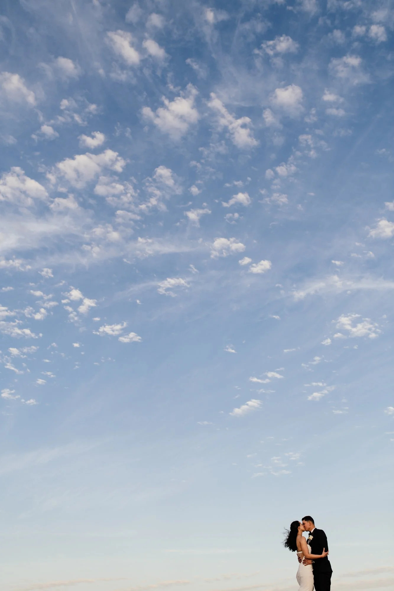A couple in wedding attire embracing and kissing outdoors under a blue sky with scattered clouds.