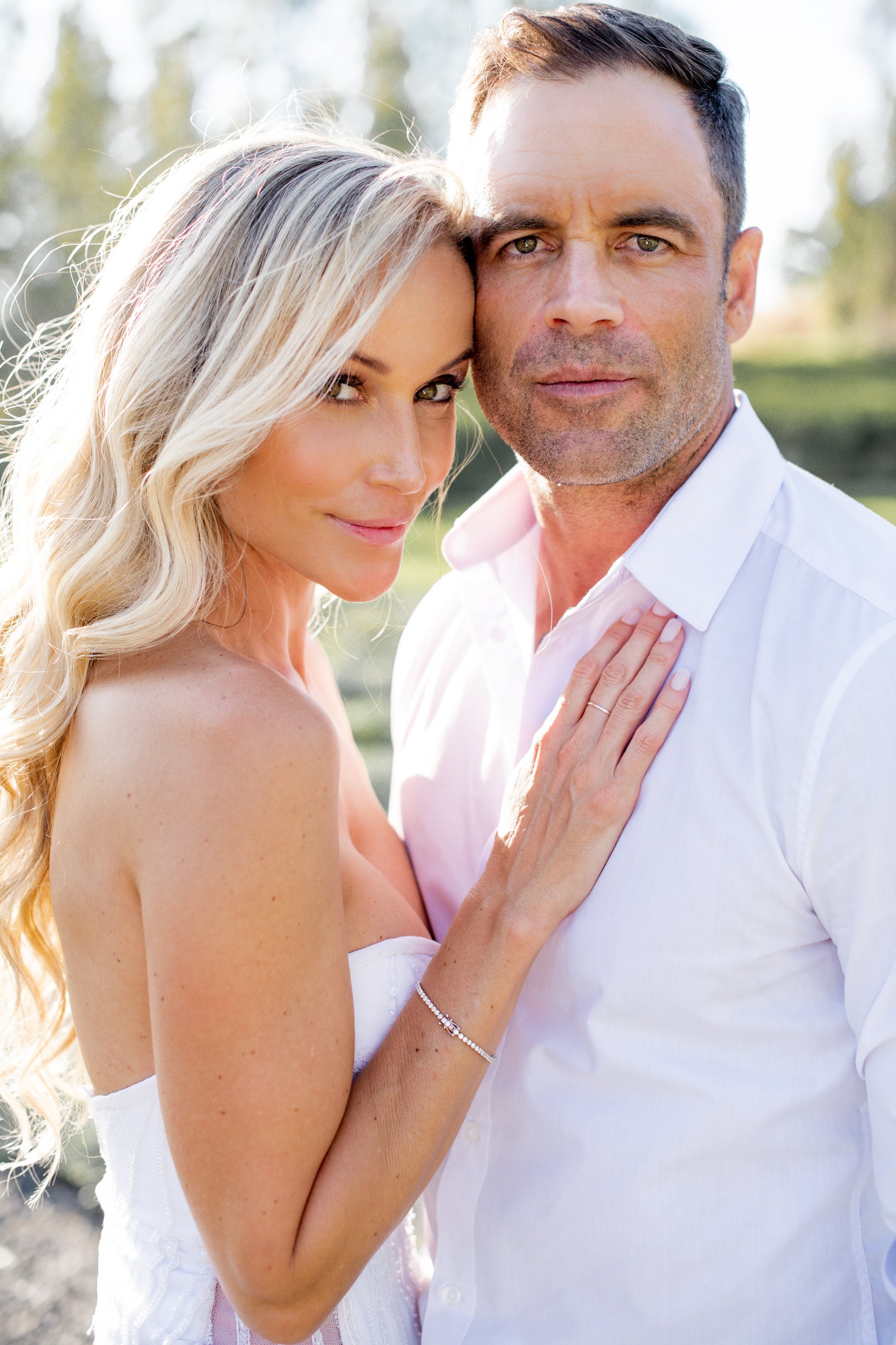 A close-up photo of a couple outdoors, with the woman leaning her head on the man's shoulder. The woman has long, wavy blonde hair and is wearing makeup, while the man has short dark hair and some facial stubble. The woman is gently touching the man’
