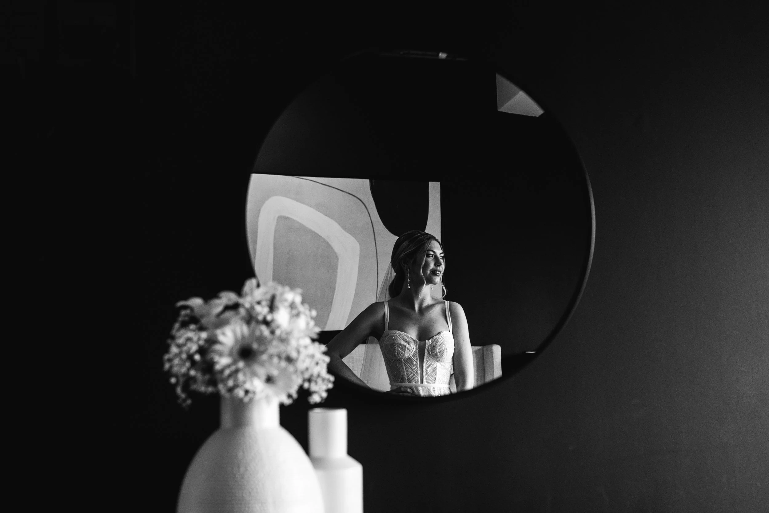 A black and white photo of a woman sitting at a table with flowers in front of her, seen through a circular mirror.
