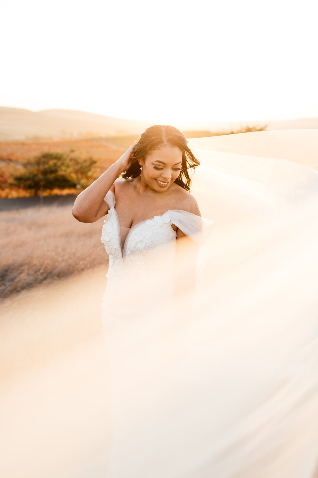 A woman in a white off-the-shoulder dress smiling outdoors during sunset, with a blurred landscape of hills and trees in the background.