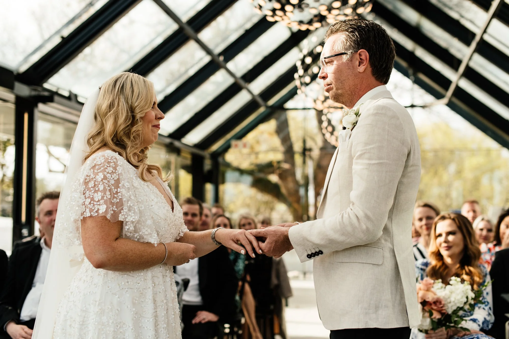 A bride and groom holding hands during their wedding ceremony, facing each other under a glass ceiling with string lights. The bride is wearing a white lace wedding dress, and the groom is dressed in a light-colored suit with glasses.