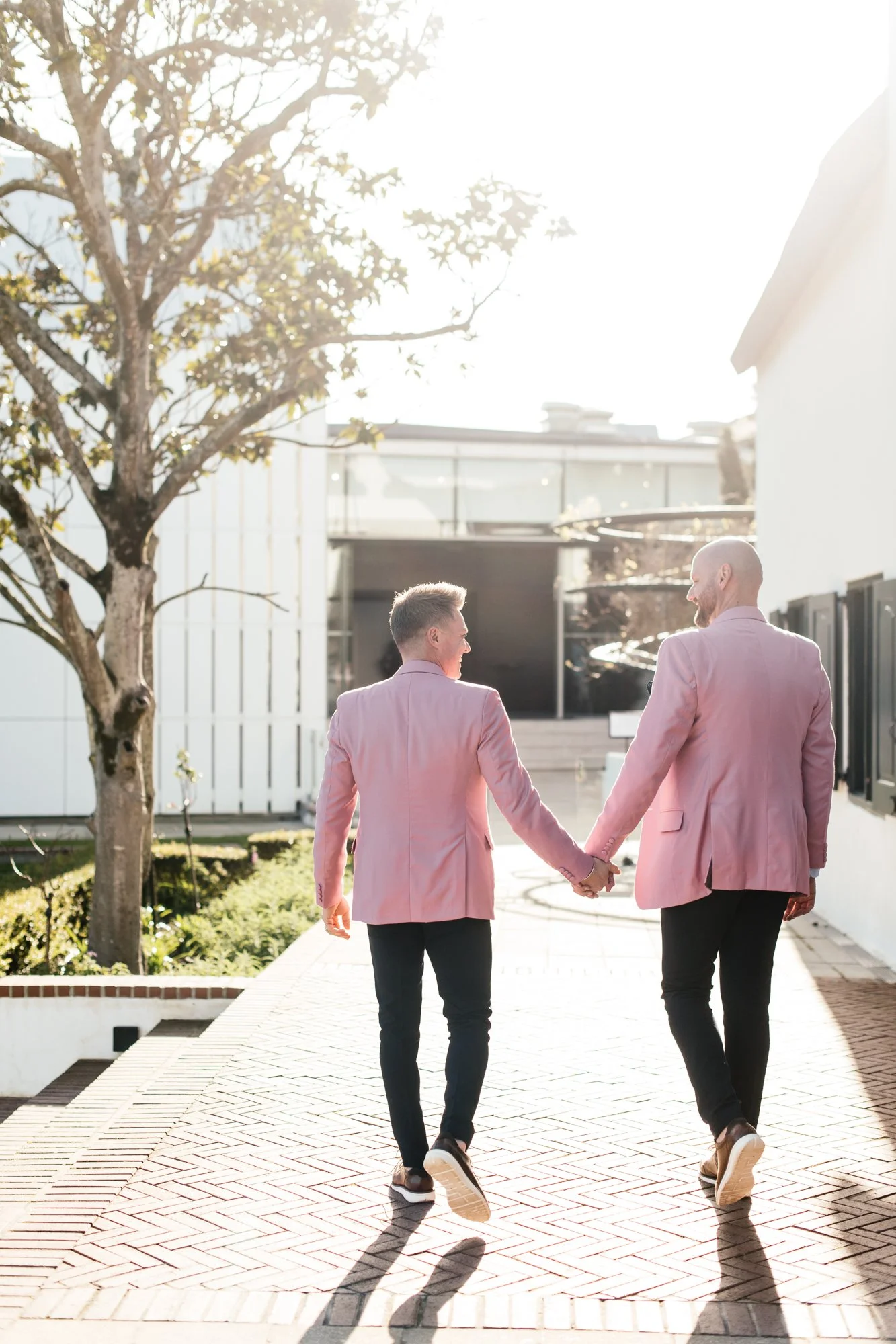 Two men walking hand in hand outdoors on a sunny day, wearing pink blazers and black pants, with buildings and trees in the background.