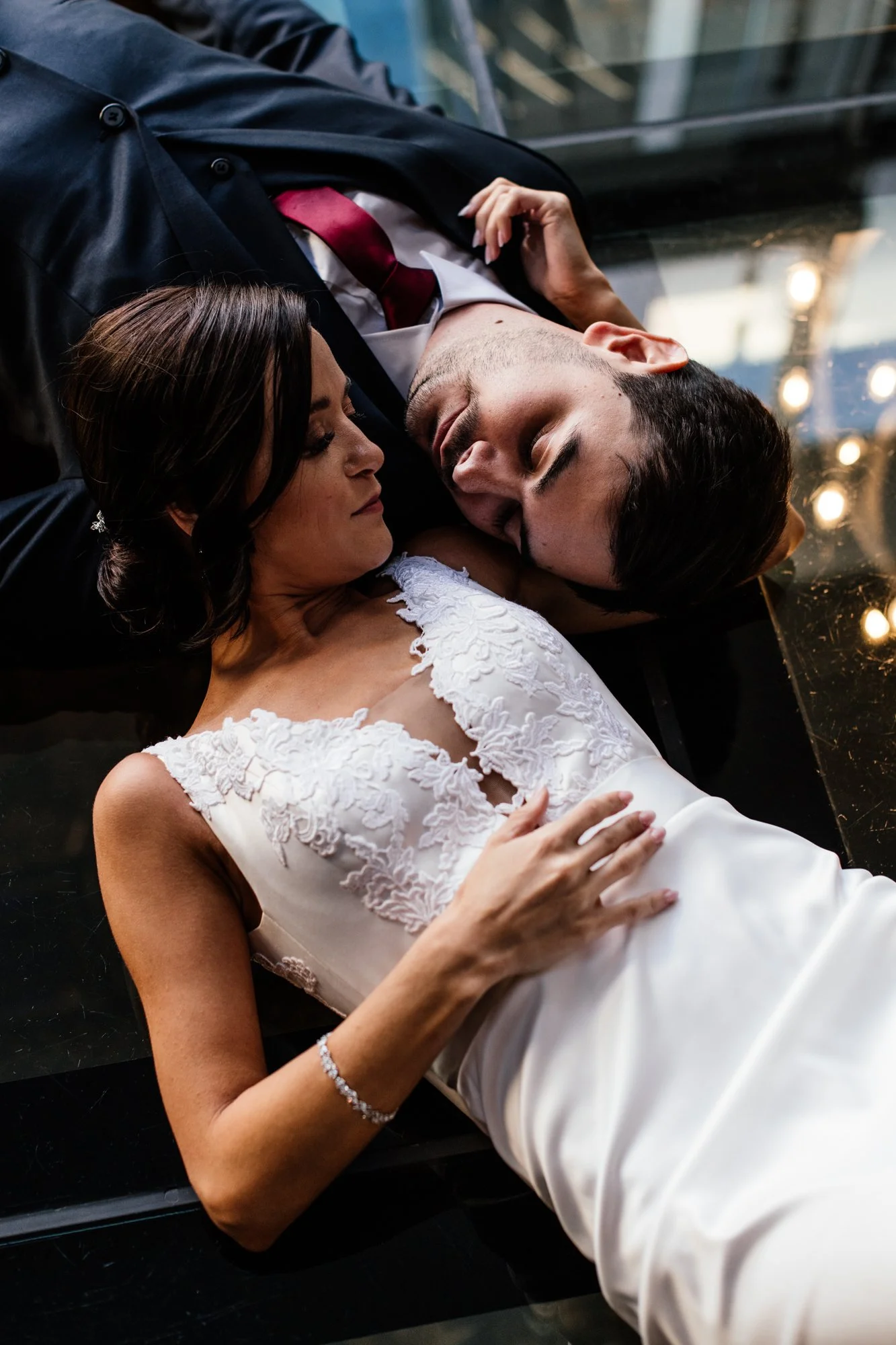 A bride and groom lying close to each other on a reflective surface, with the bride wearing a white lace wedding dress and the groom in a dark suit with a red tie, sharing an intimate moment.