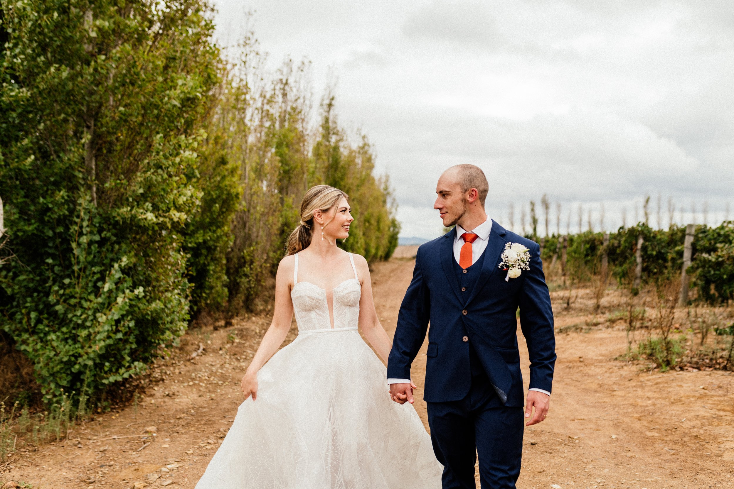 A bride and groom happily holding hands and walking outdoors in a vineyard on a cloudy day, smiling at each other.