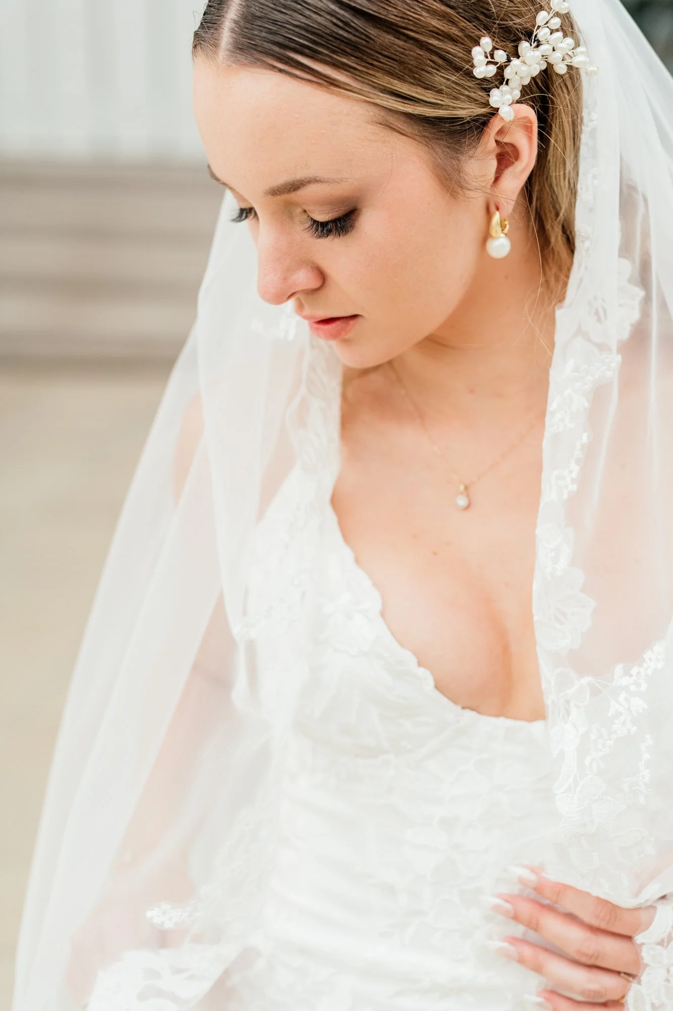 A bride with princess-like makeup and earrings, wearing a white lace wedding gown, and a sheer veil, posing with her head slightly tilted downward and her hand resting on her waist.