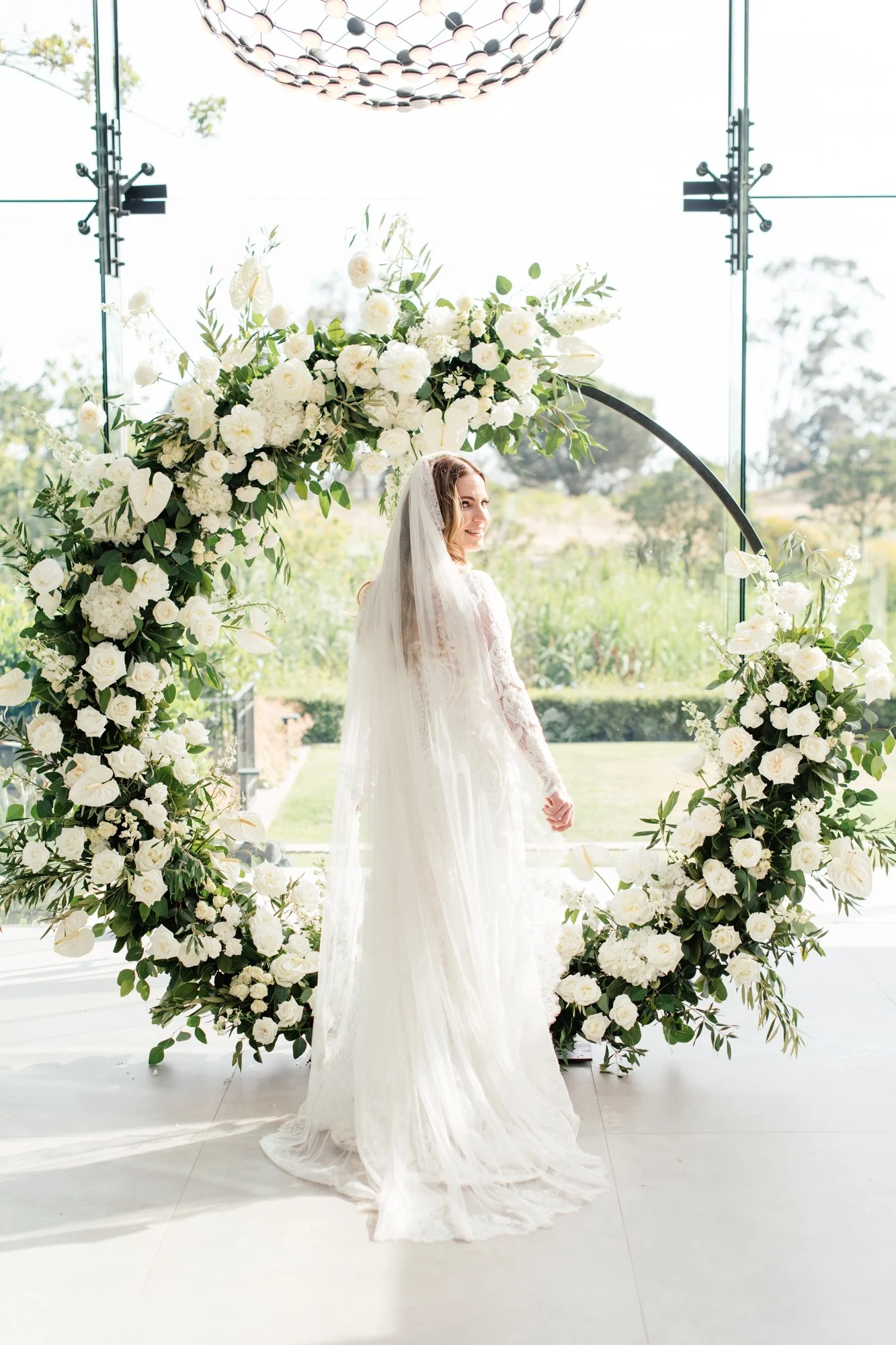 A woman in a white wedding dress with long lace sleeves and a veil standing outdoors beneath a circular floral arch of white flowers and greenery, during daytime with trees and a glass structure background.