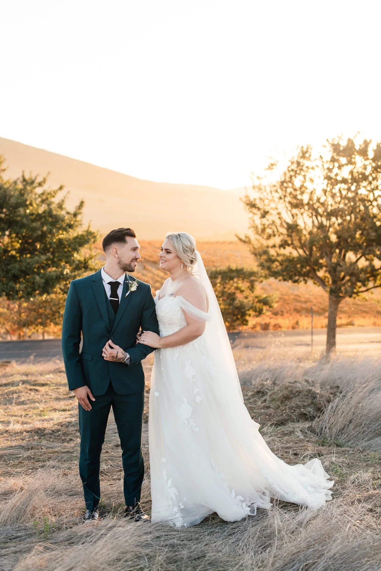 A bride and groom stand outdoors during sunset, holding hands and gazing at each other, with a mountain and trees in the background.