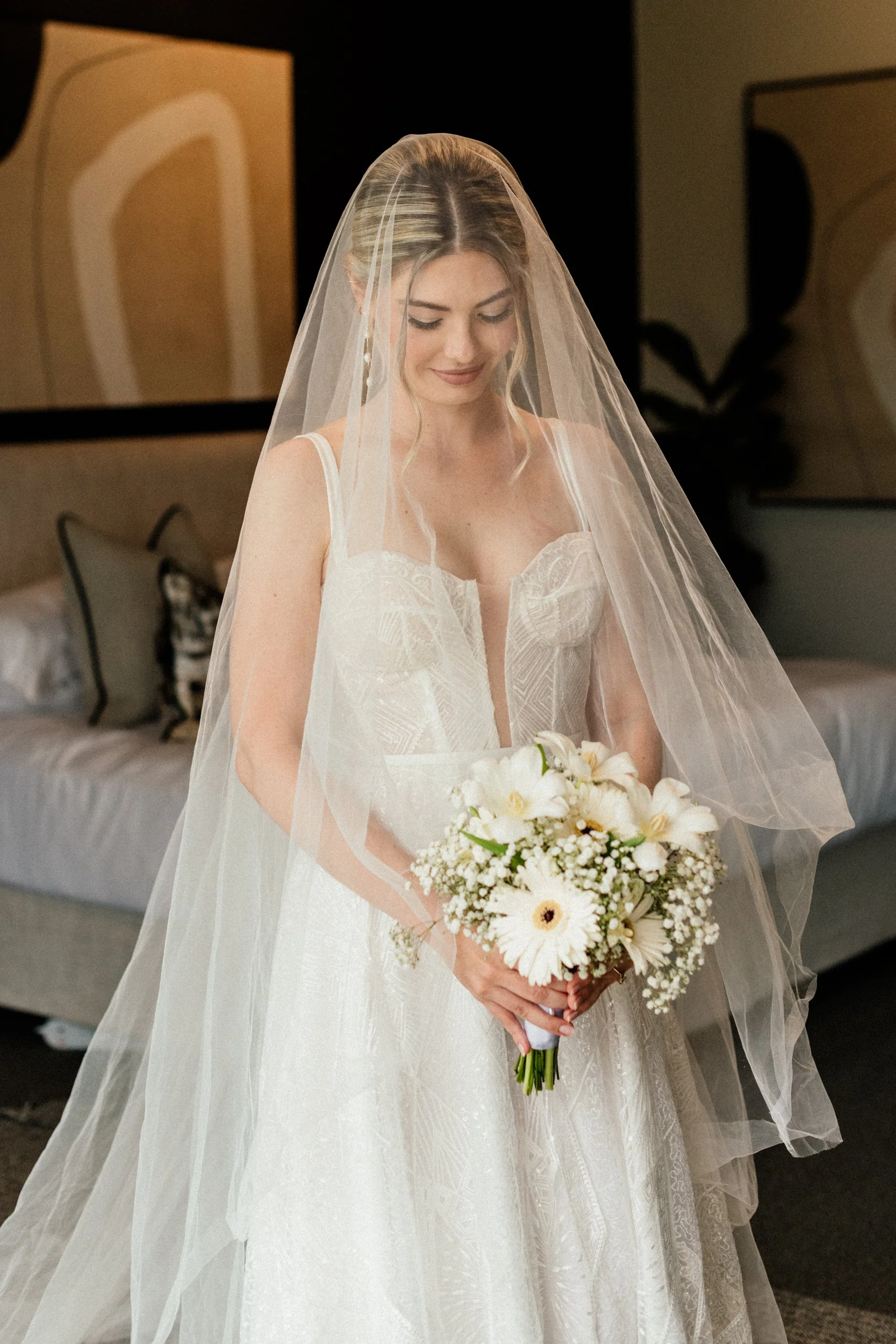 A bride with blonde hair and a white wedding dress, holding a bouquet of white flowers, and wearing a veil, standing indoors.