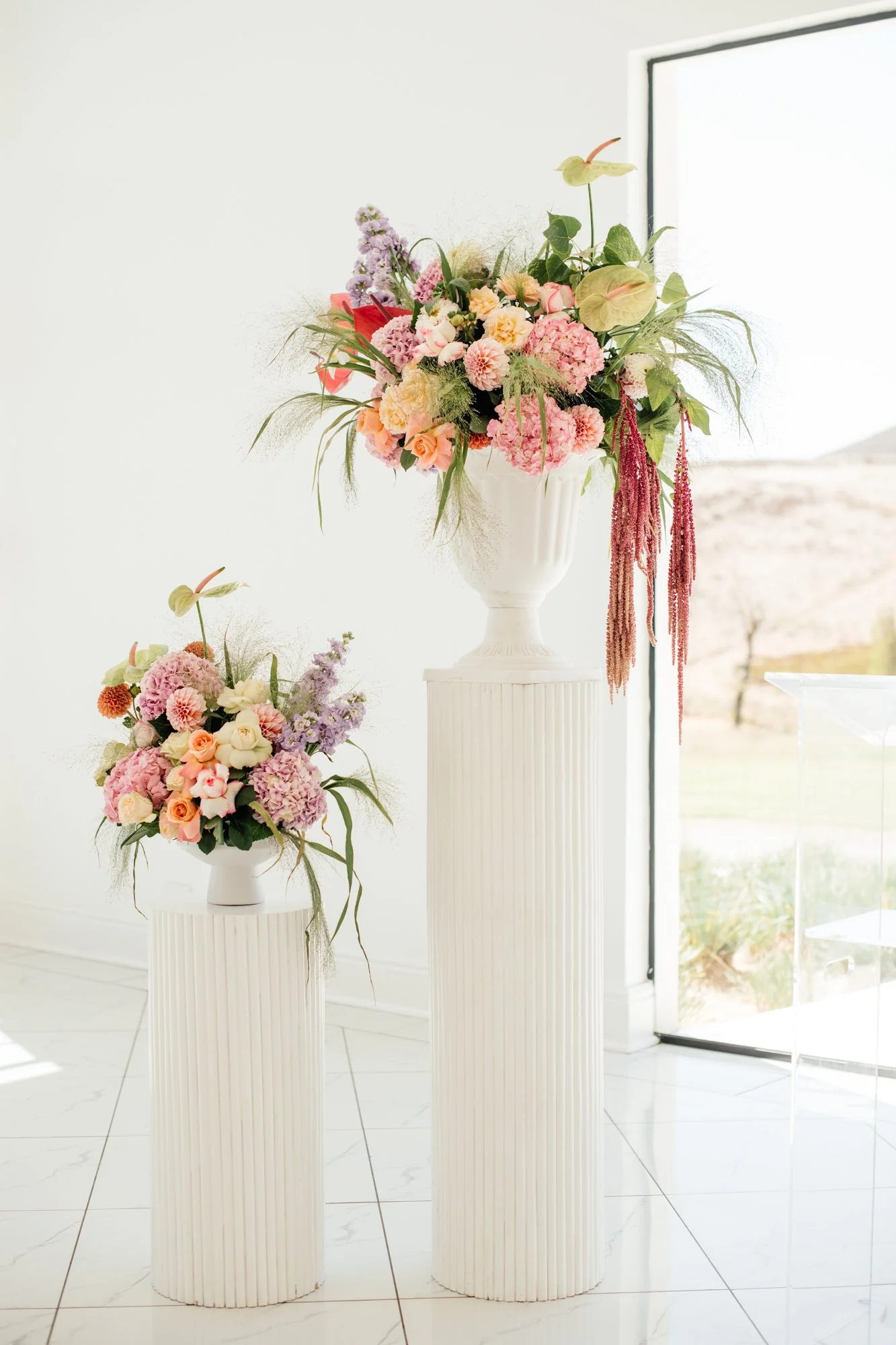 Two white vases with pink and purple floral arrangements are placed on white fluted pedestals inside a bright room with white walls and tiles. A window on the right shows an outdoor landscape with fields and trees.