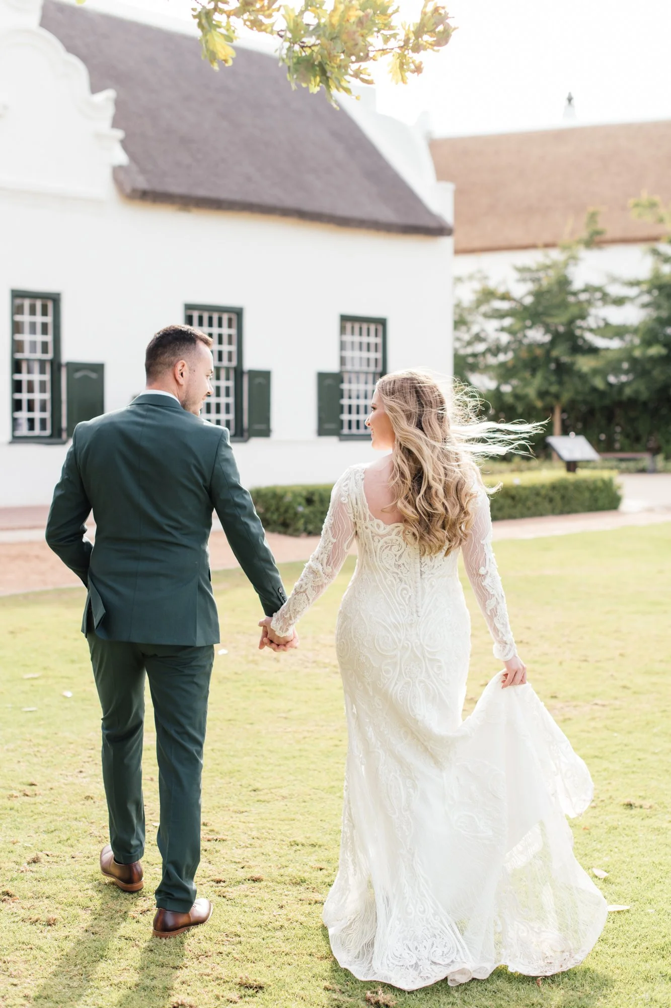 A newlywed couple holding hands and walking on a grassy lawn in front of a white building with dark window frames, during daytime.