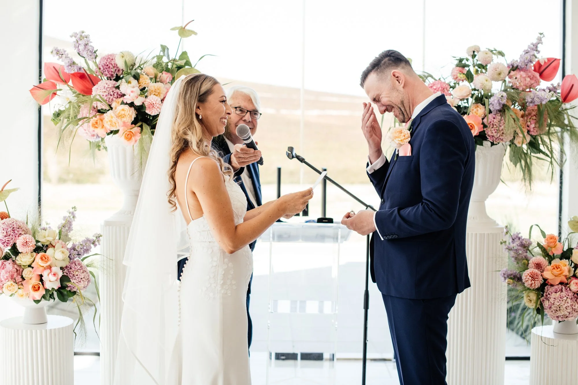 A bride and groom exchanging vows during a wedding ceremony with floral arrangements in the background.