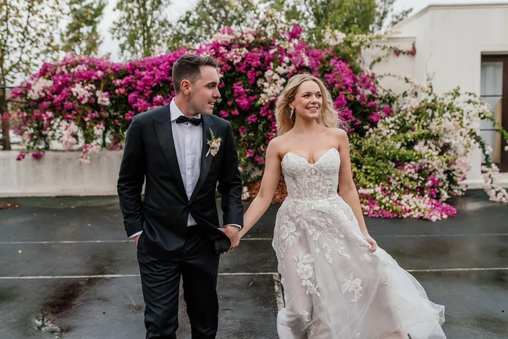 Bride and groom holding hands outdoors with vibrant pink bougainvillea flowers in the background.