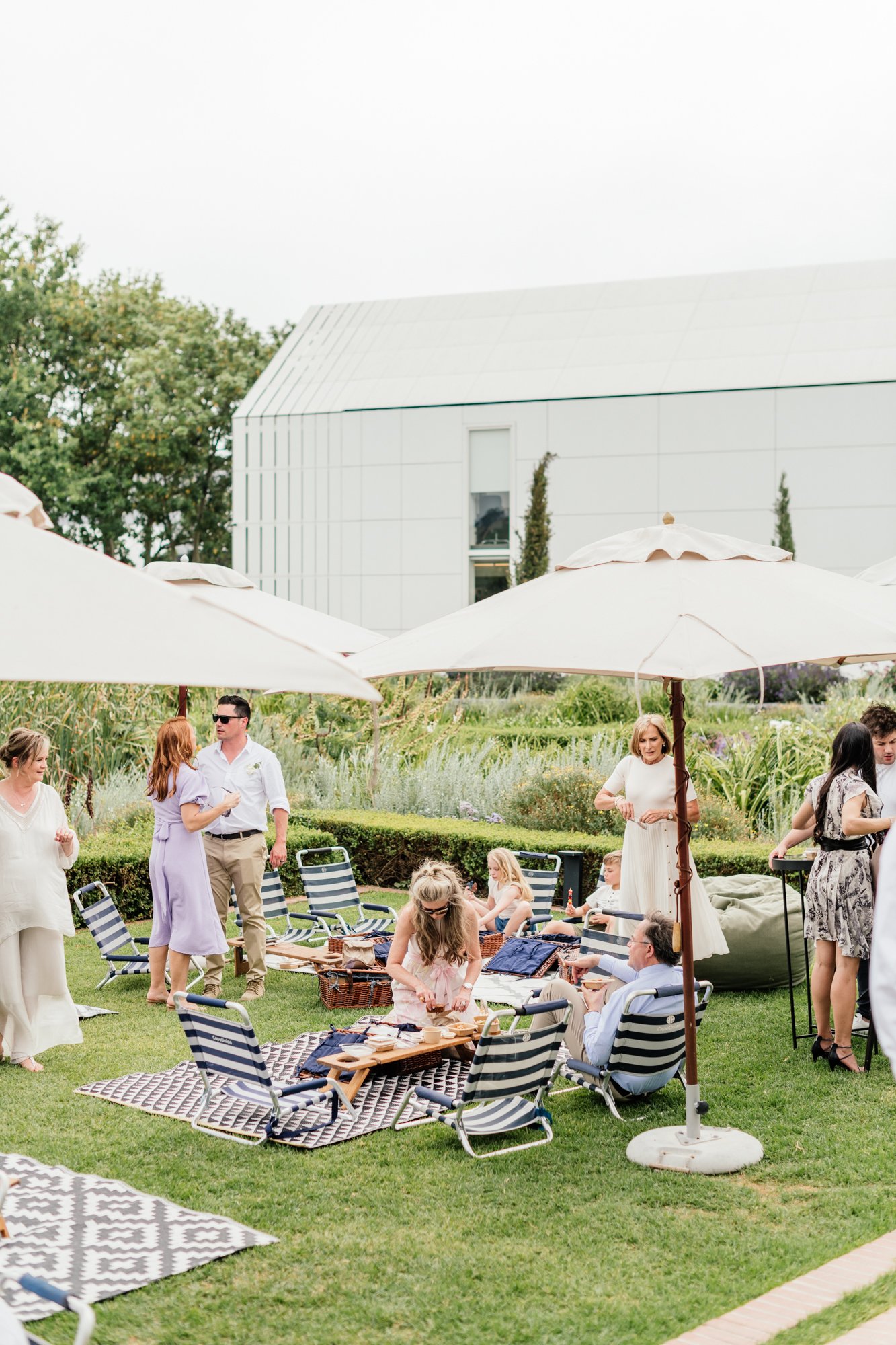 People enjoying a daytime outdoor gathering in a garden with umbrellas, chairs, and picnic tables, with a modern building and trees in the background.