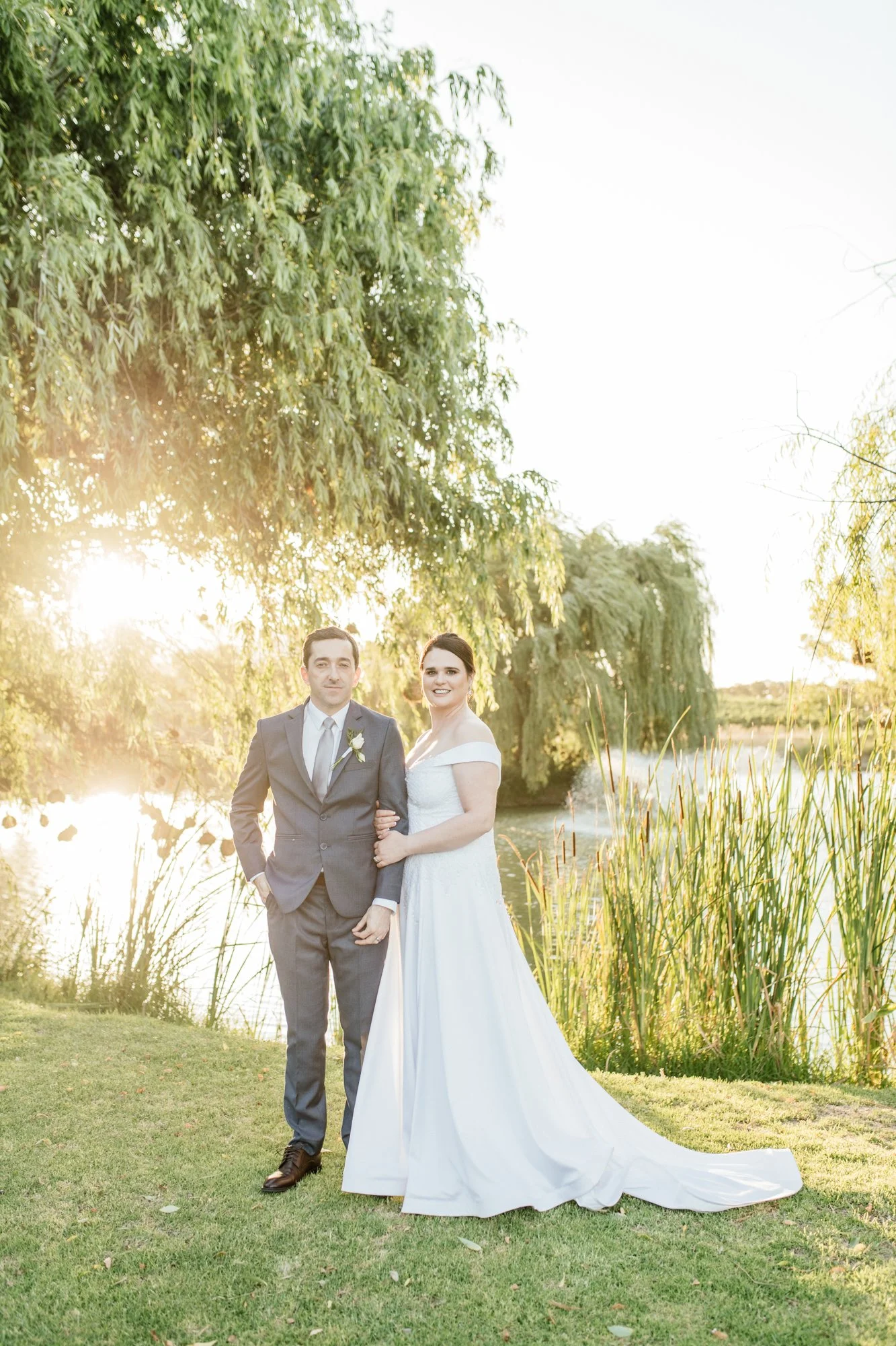 A married couple in wedding attire standing on grass near a lake, with tall trees and reeds in the background, during sunset.