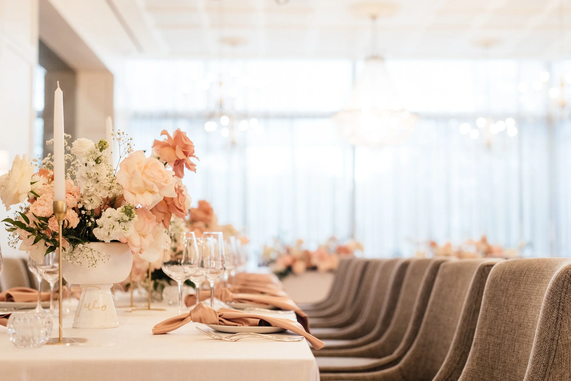 Elegant banquet table set for a wedding or special event, decorated with a large floral centerpiece, candlesticks, glassware, and napkins, with a bright, airy background featuring large windows and ceiling chandeliers.
