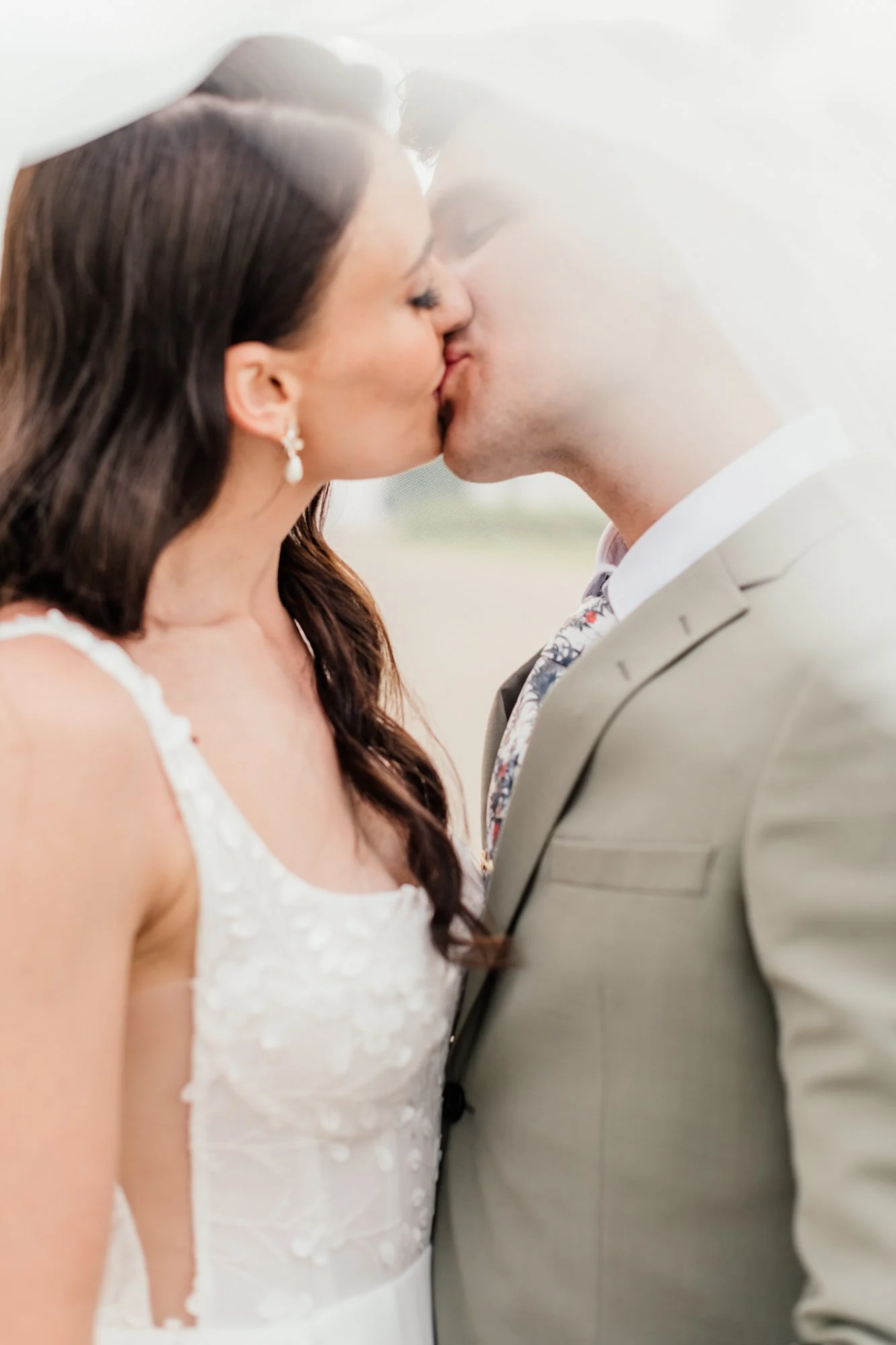 A bride and groom kissing, seen through a foggy or frosted glass. The bride has long dark hair, pearl earrings, and wears a white wedding dress with lace details. The groom is wearing a light-colored suit and a patterned tie.