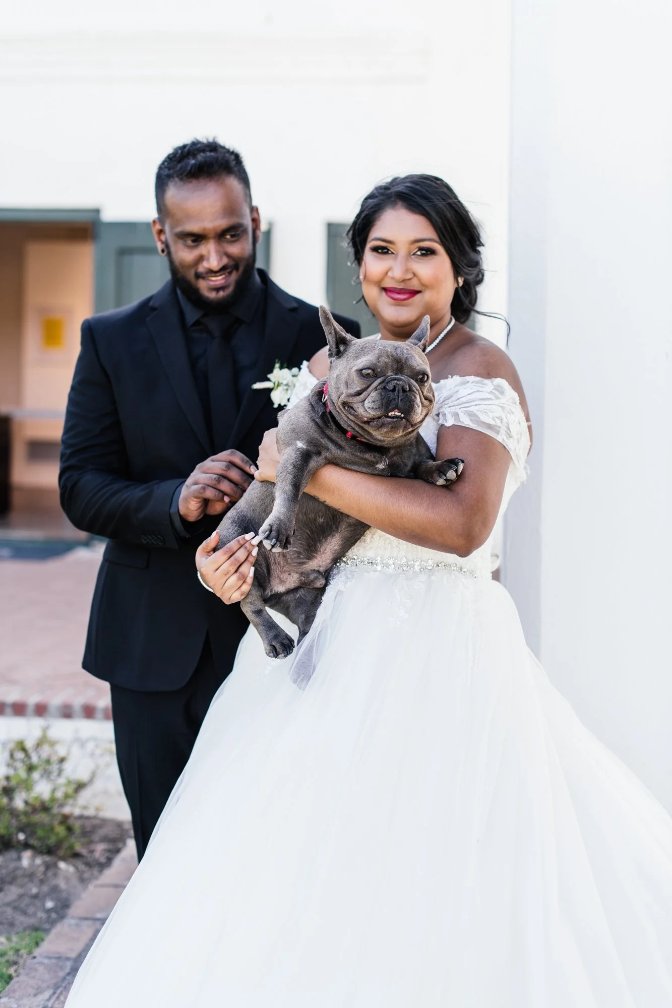 A bride and groom in wedding attire holding a happy French Bulldog dog outside near a white wall and garden.
