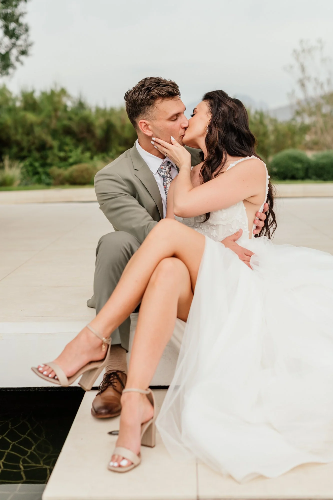 A man and woman in wedding attire sharing a kiss, with the woman sitting on the man’s lap outdoors on a patio with greenery and overcast sky in the background.