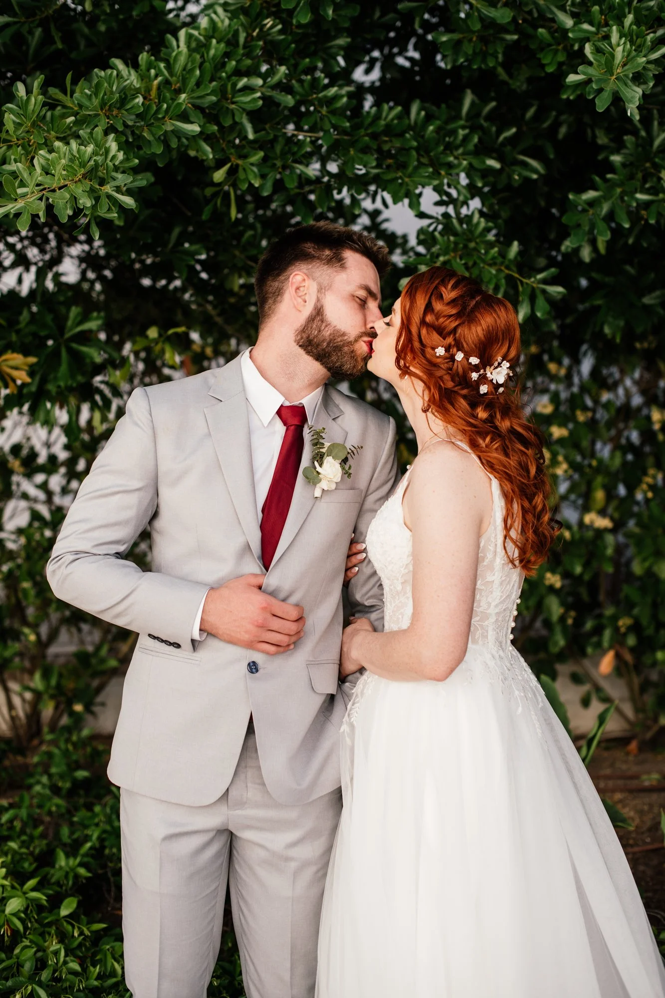 A couple in wedding attire kissing outdoors in front of green foliage.