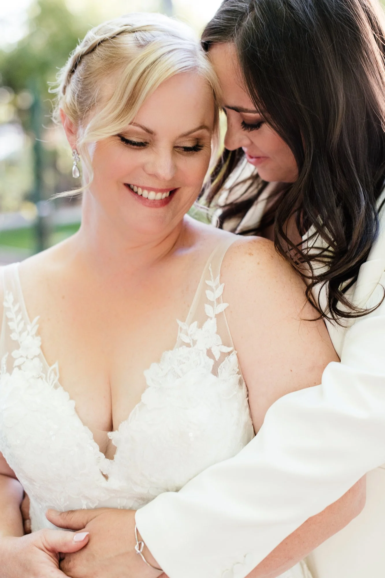 Two women, one in a wedding dress and the other in a white dress, sharing a close, joyful moment outdoors. LBGTQ+ Gay friendly.
